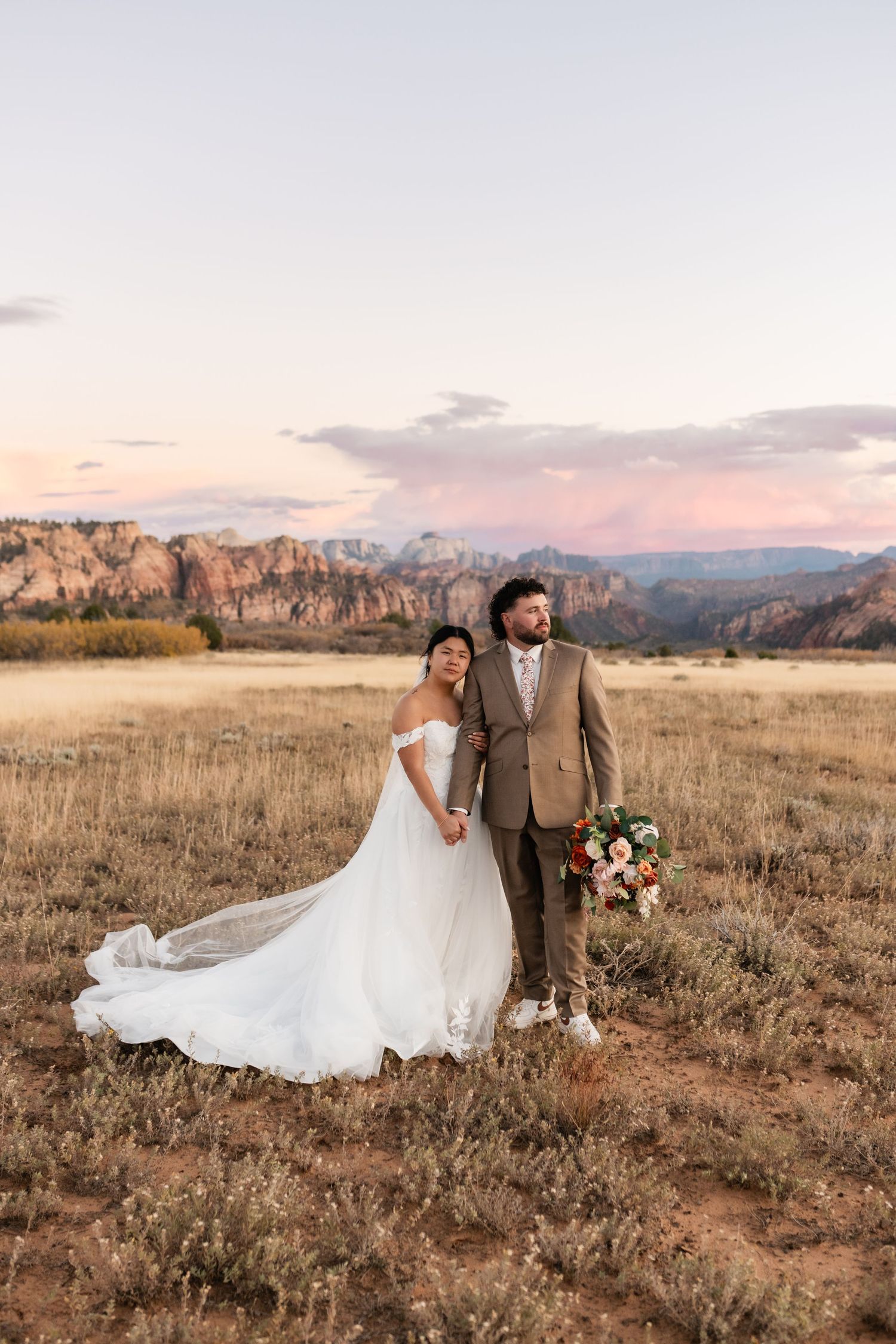 Bride and groom pose in desert field during sunset with red rock cliffs in background.