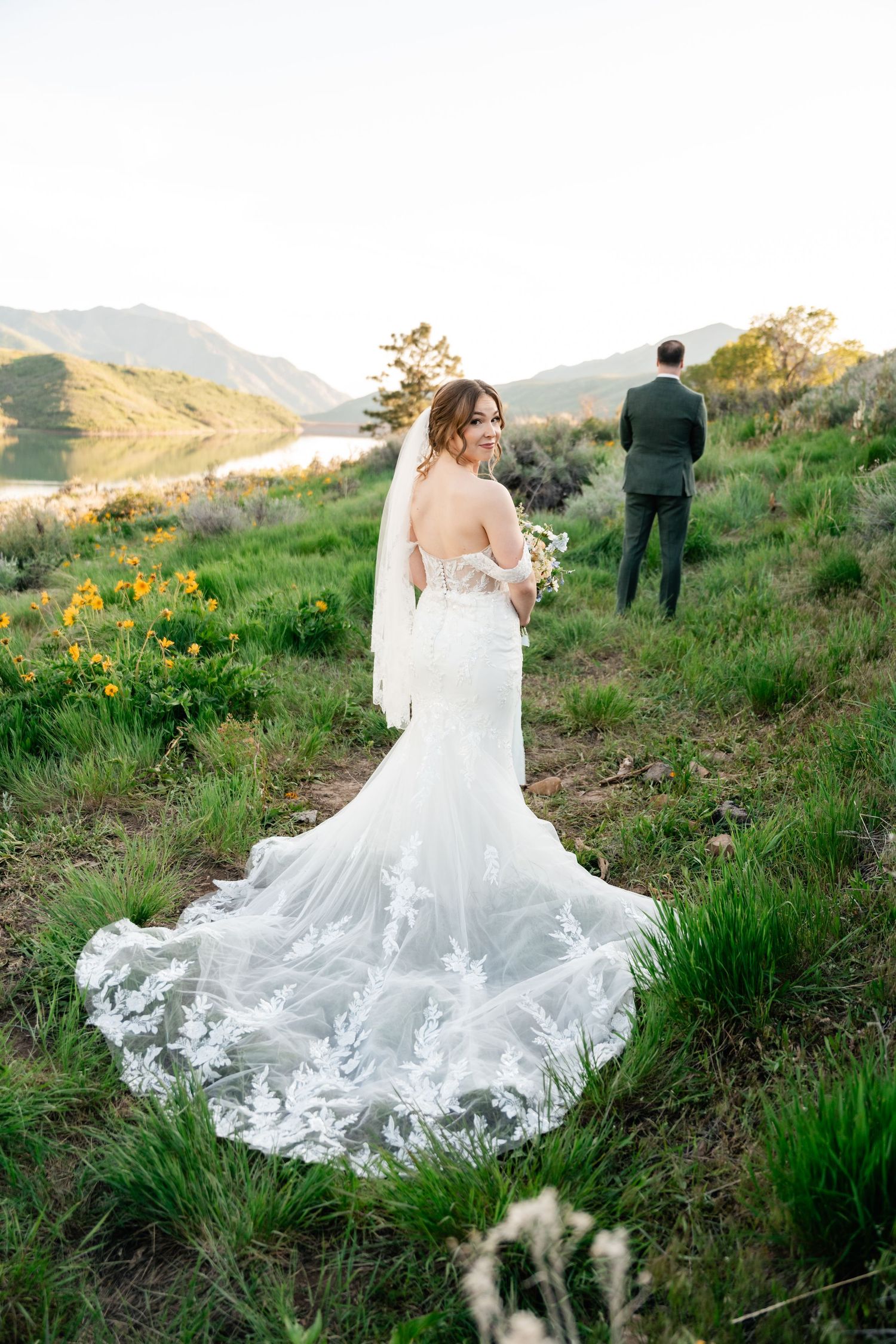 Elegant white wedding dress flowing across a hillside meadow with yellow wildflowers and mountains in the background.