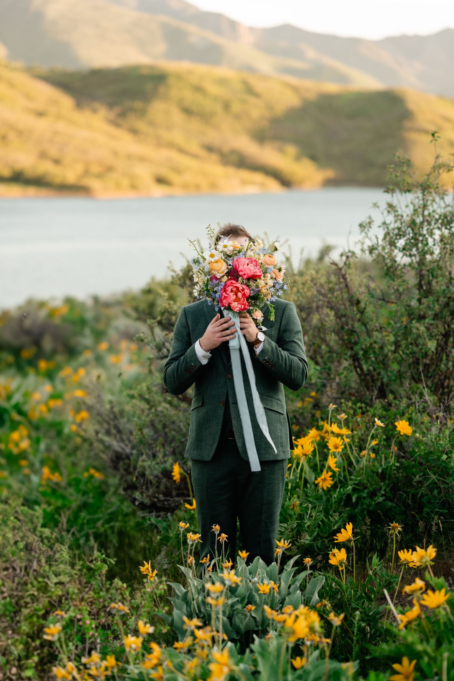 Someone in a green suit holds a vibrant bouquet while standing among yellow wildflowers on a hillside by a lake.