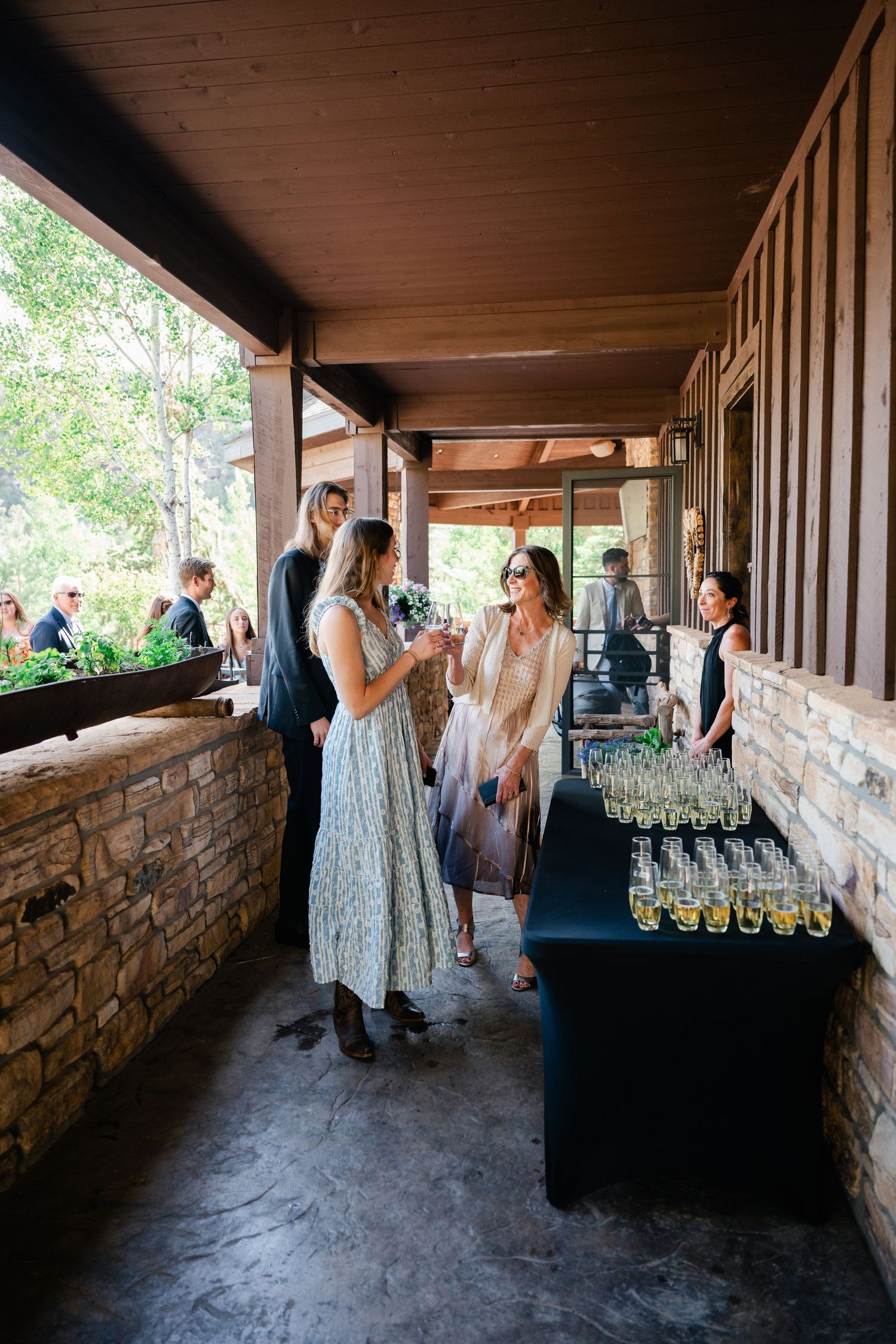 Wedding guests mingle and socialize in a rustic covered patio area with stone walls.