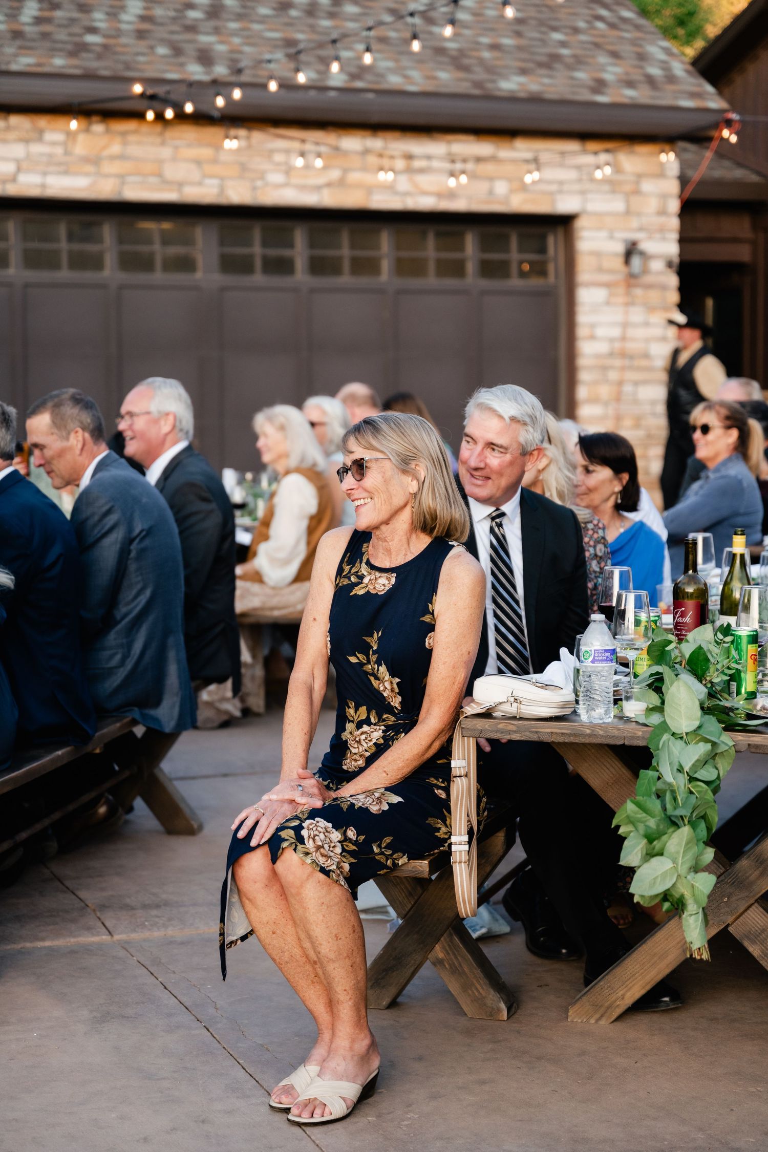 Wedding guests relax at rustic wooden picnic tables under string lights during an outdoor reception.