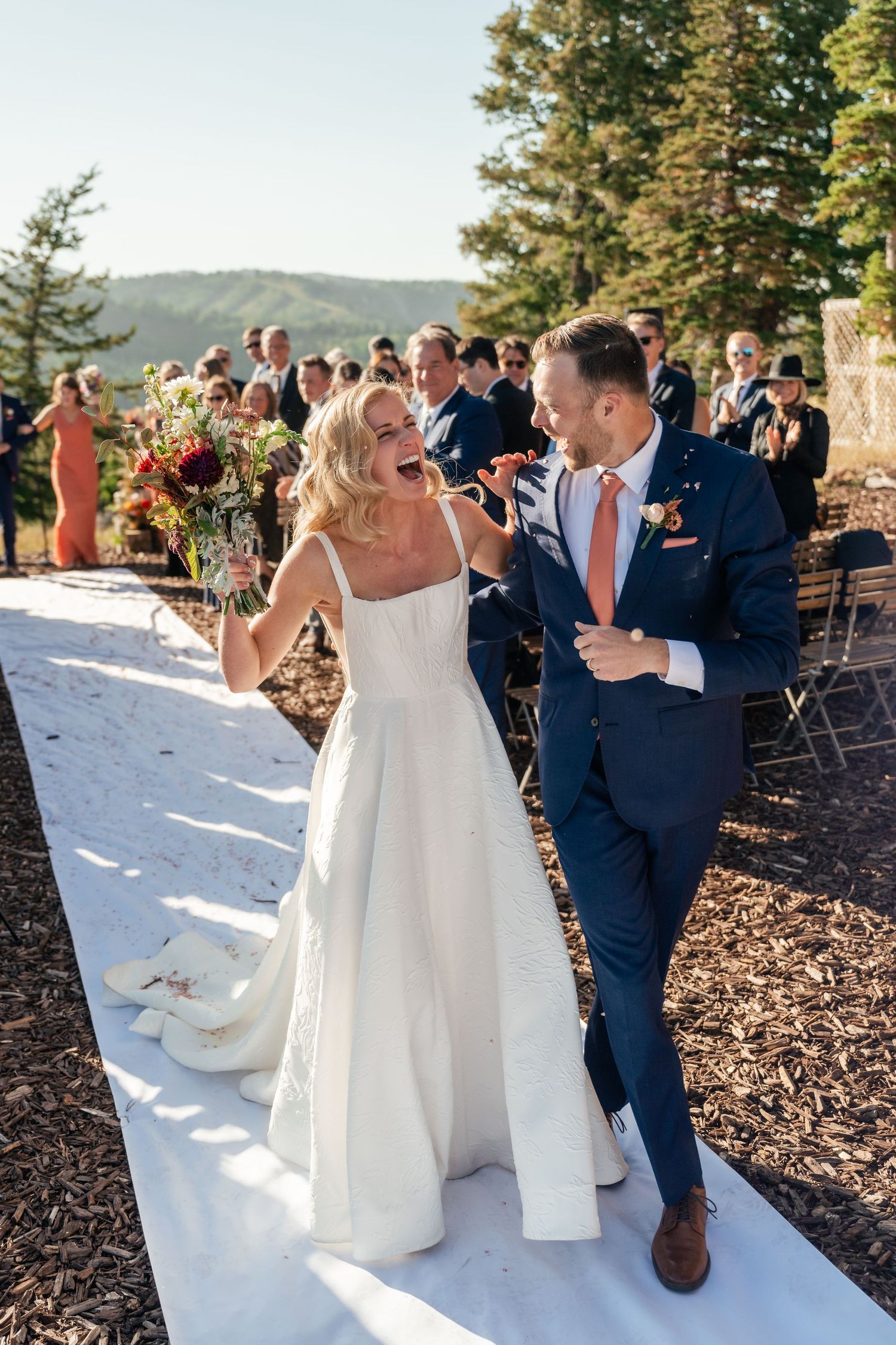 Wedding couple walking down white aisle runner outdoors with mountain scenery in background.