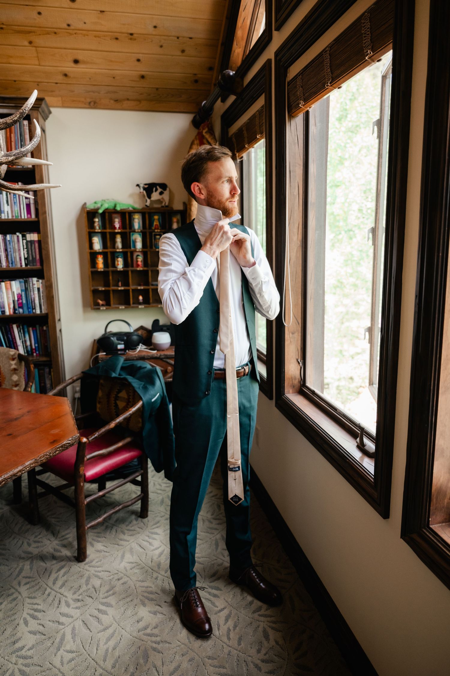 Groom in green vest looks out window while getting ready in rustic cabin setting.