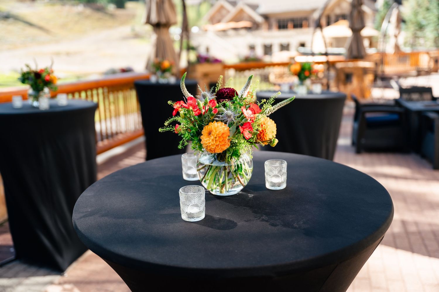 Small floral centerpieces with orange and pink blooms sit on black tablecloths at an outdoor event setting.