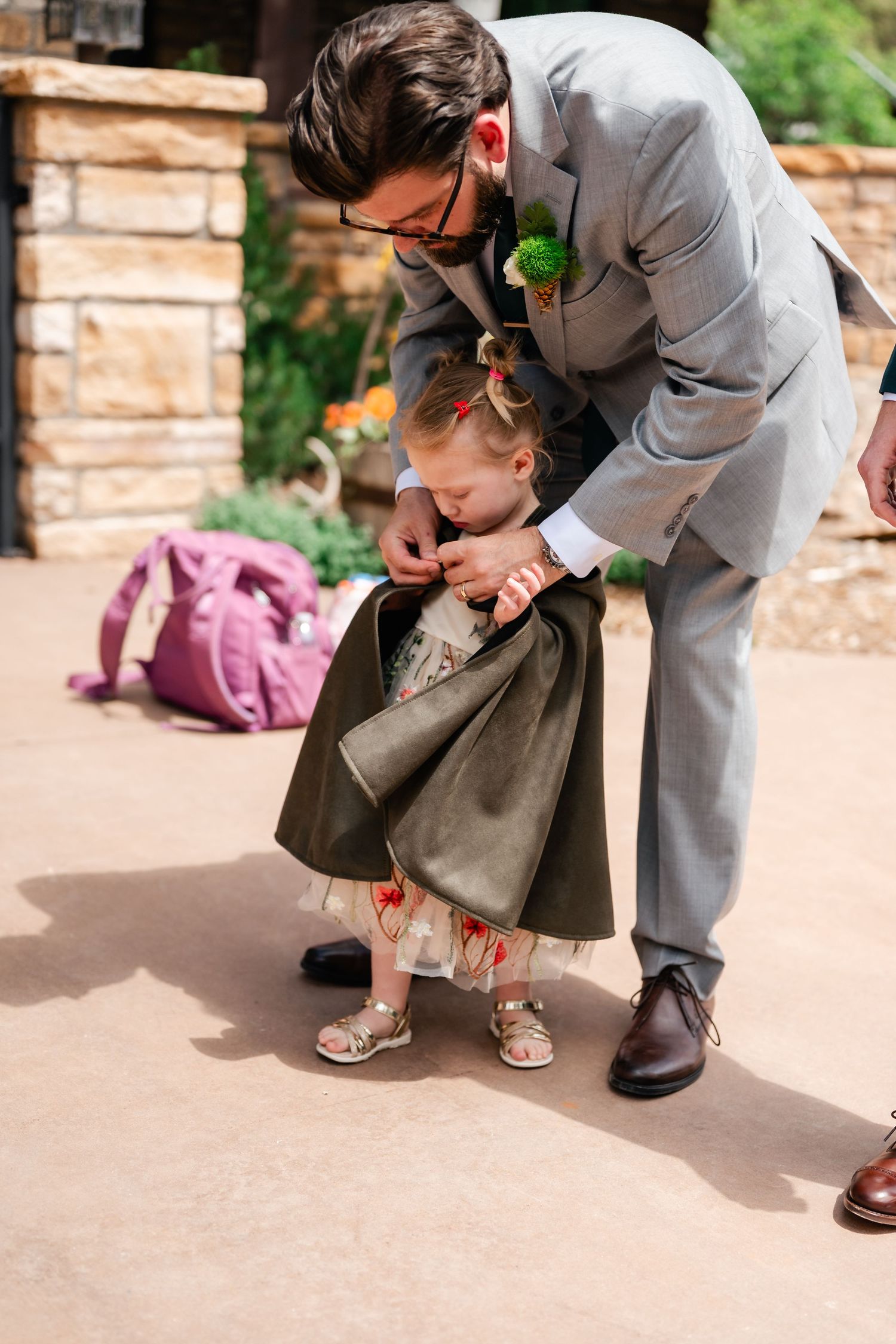 Person in gray suit kneeling to adjust child's formal attire outdoors.