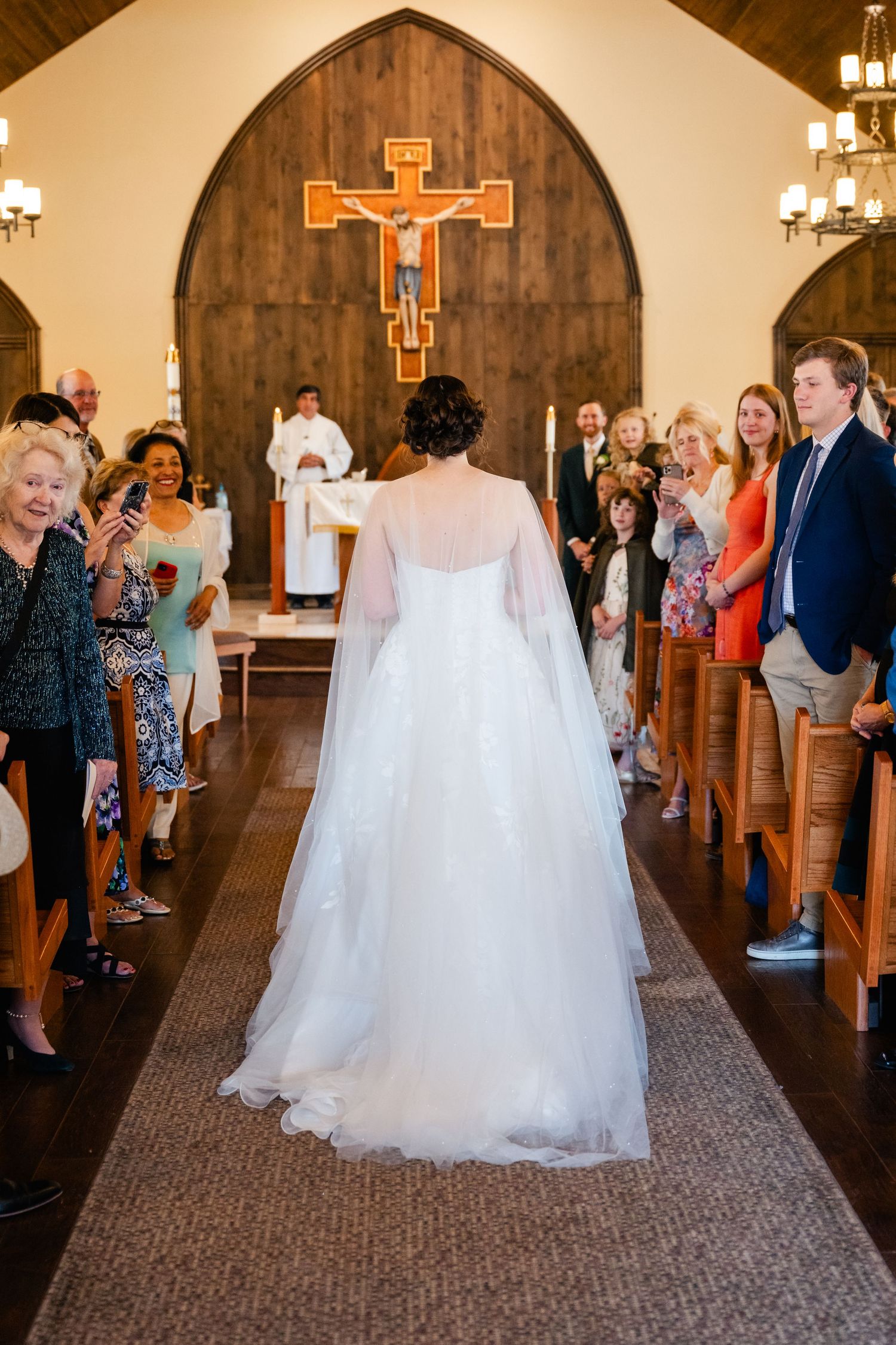 A bride in a white gown walks down the church aisle with wooden crosses on the wall.