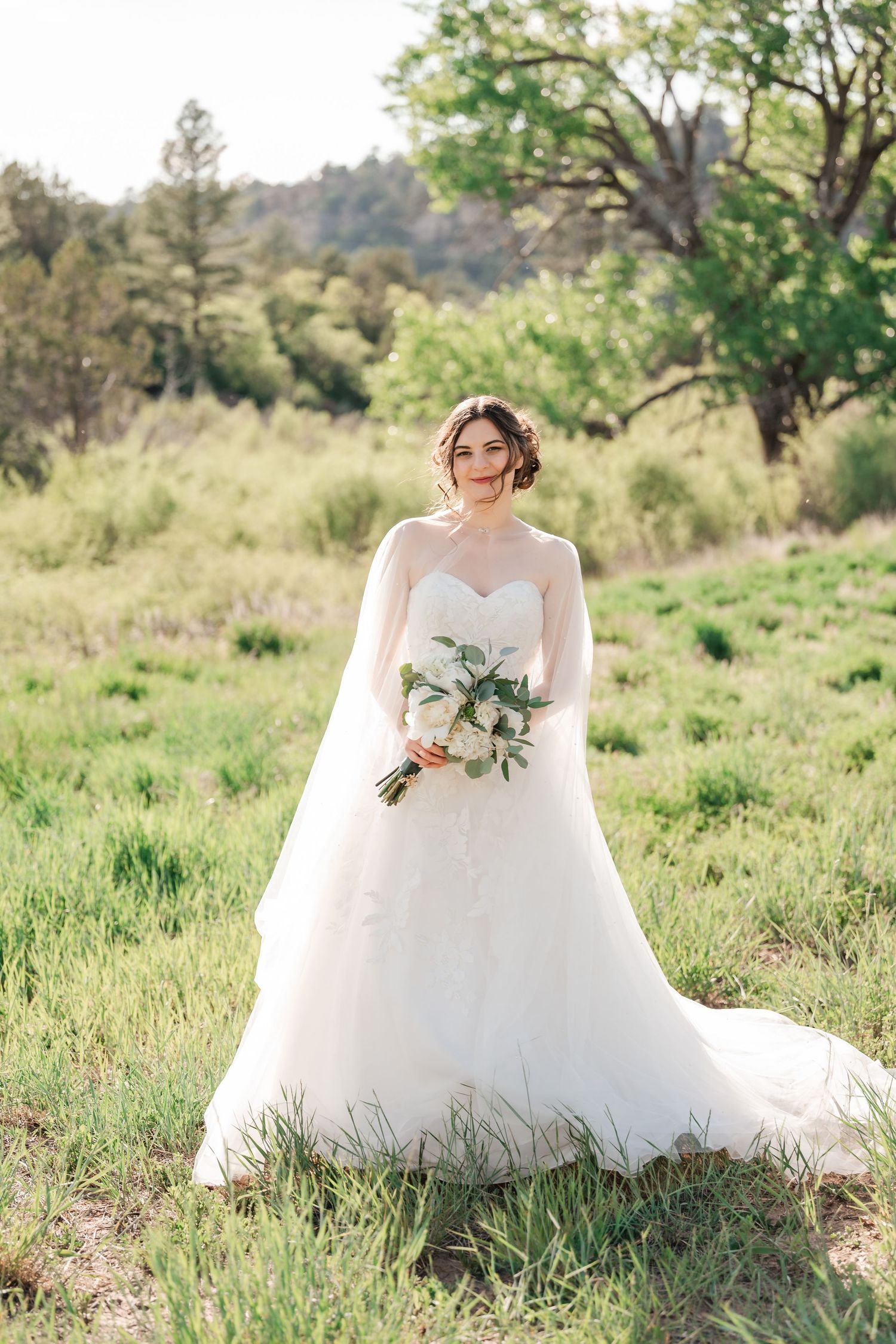 A bride in a flowing white gown poses in a sunlit meadow with spring greenery.
