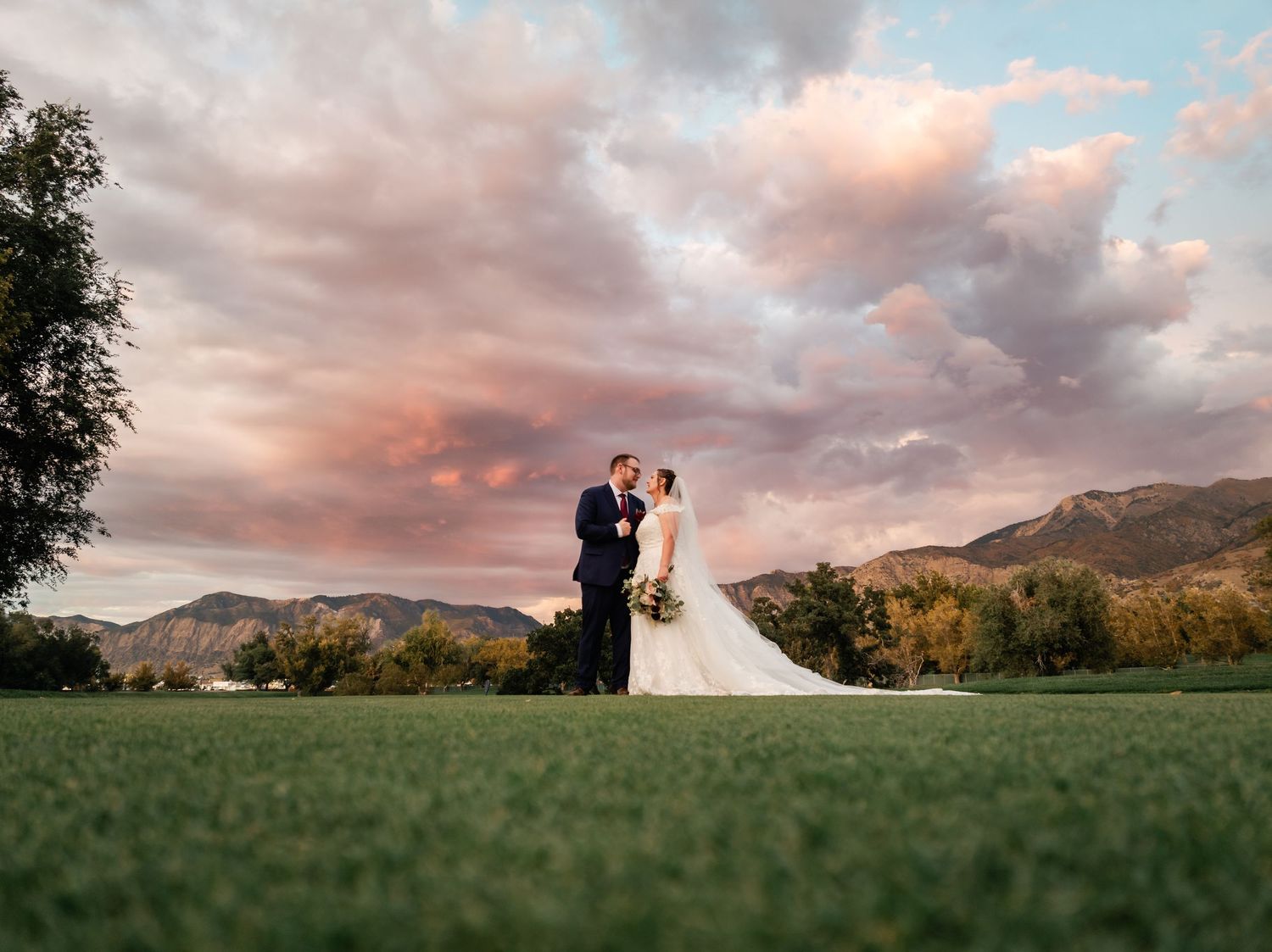 A couple in wedding attire stands in a grassy field at sunset with mountains and pink clouds in the background. Utah Wedding Photographer.