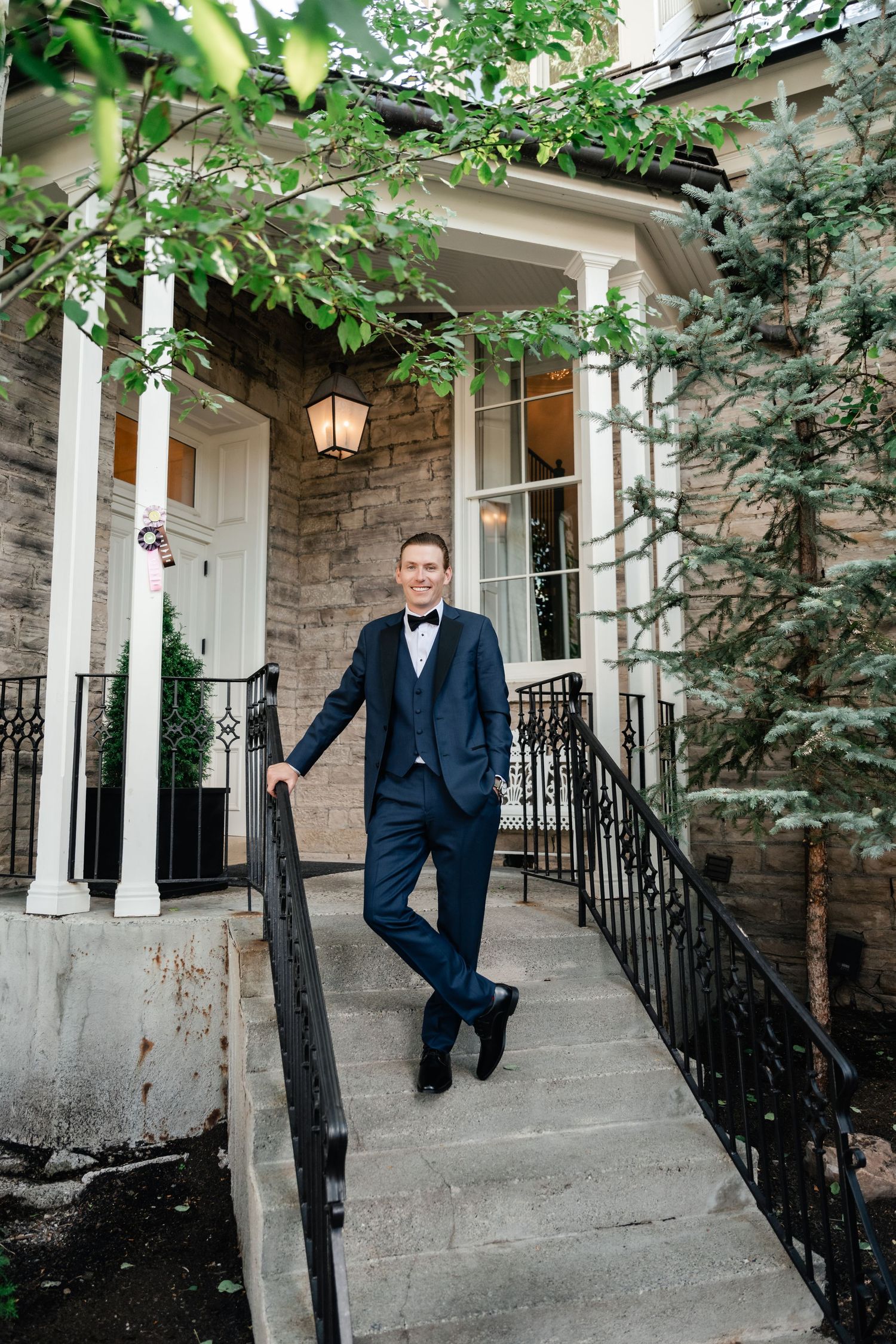 Groom in navy suit poses casually against stone building columns and iron railings.