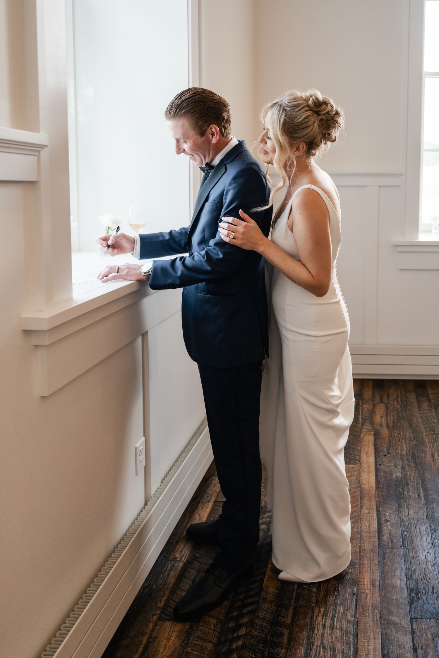 A couple in formal attire stands by a bright window writing something during a wedding celebration.