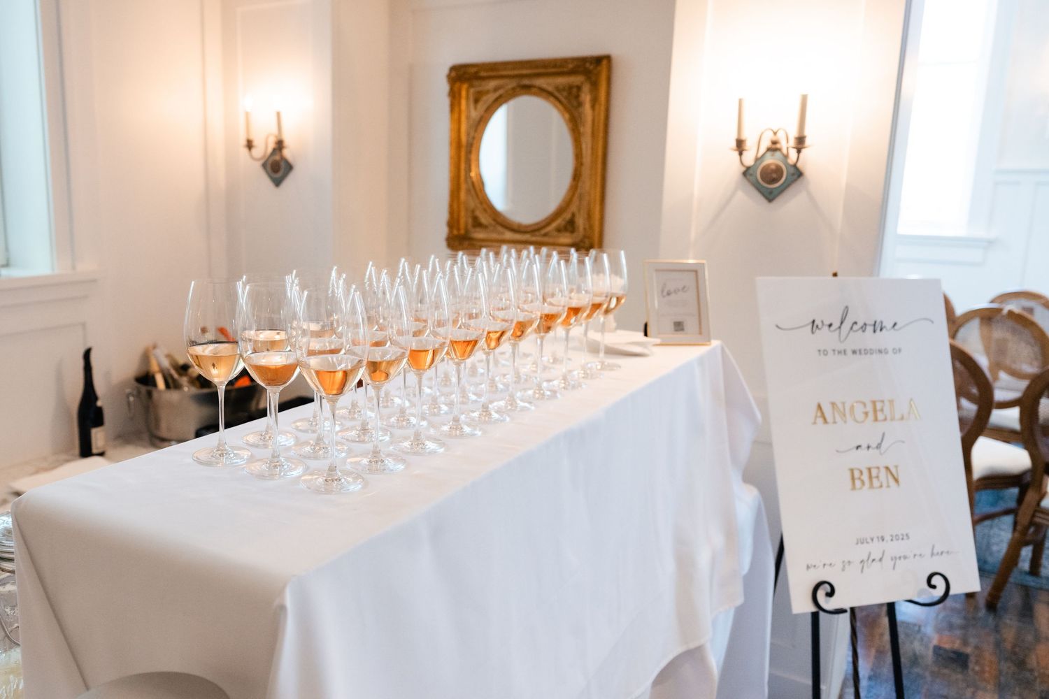 Long table with wine glasses set up for wedding reception with gold-framed mirror and welcome sign.