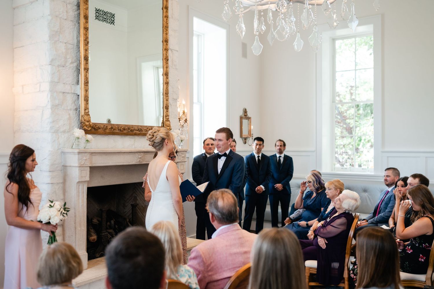 Wedding ceremony attendees gather in an elegant white room with large windows and ornate gold framing.