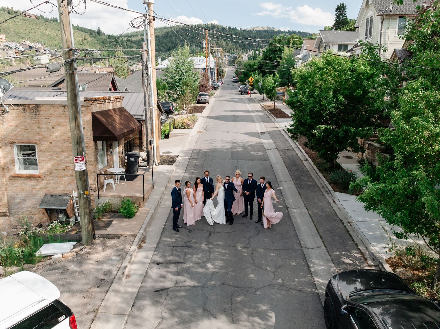 A sloped residential street lined with houses and trees showing a group of people walking in formal attire.