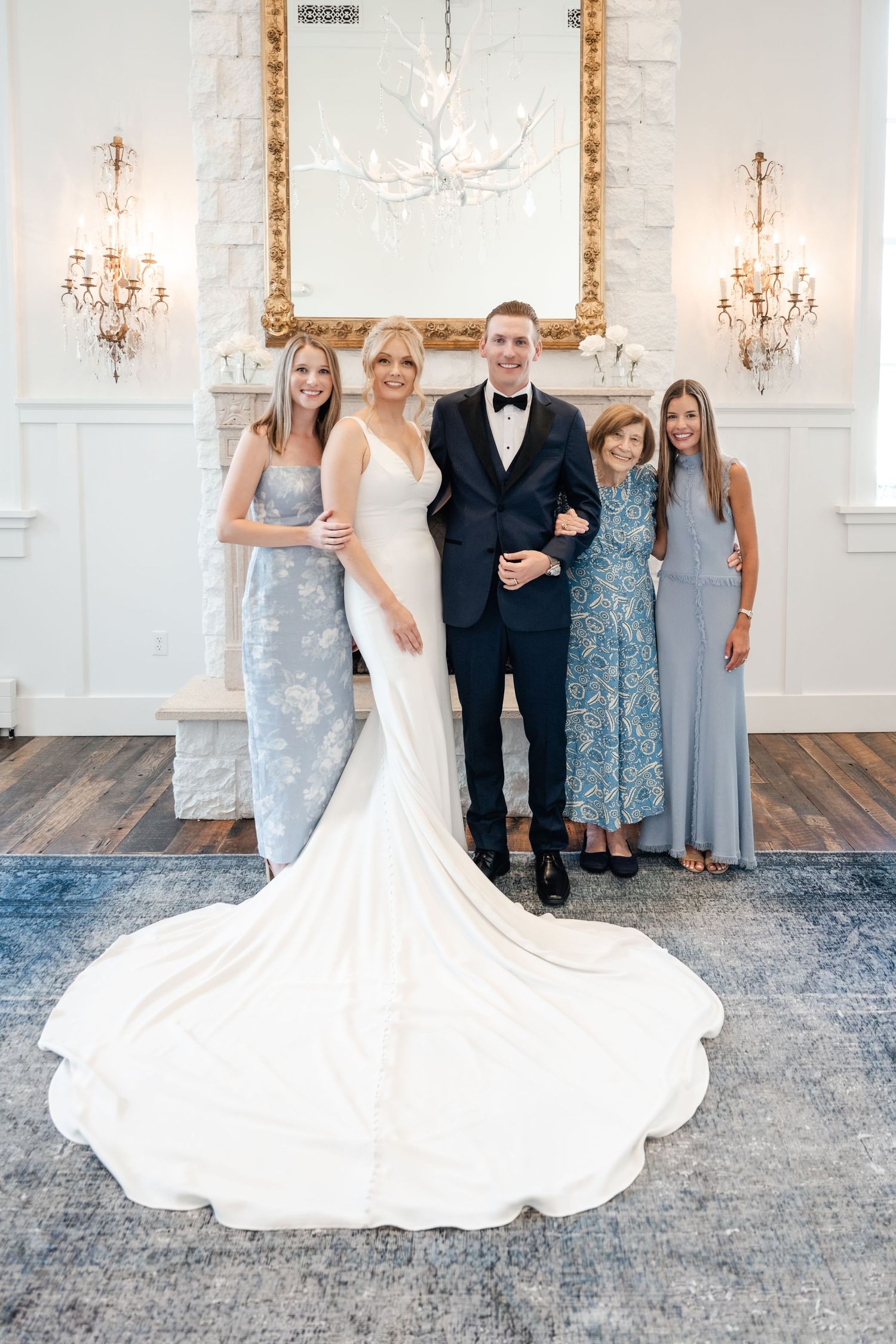 A wedding party poses in an elegant white room with crystal chandeliers and ornate wall decor.