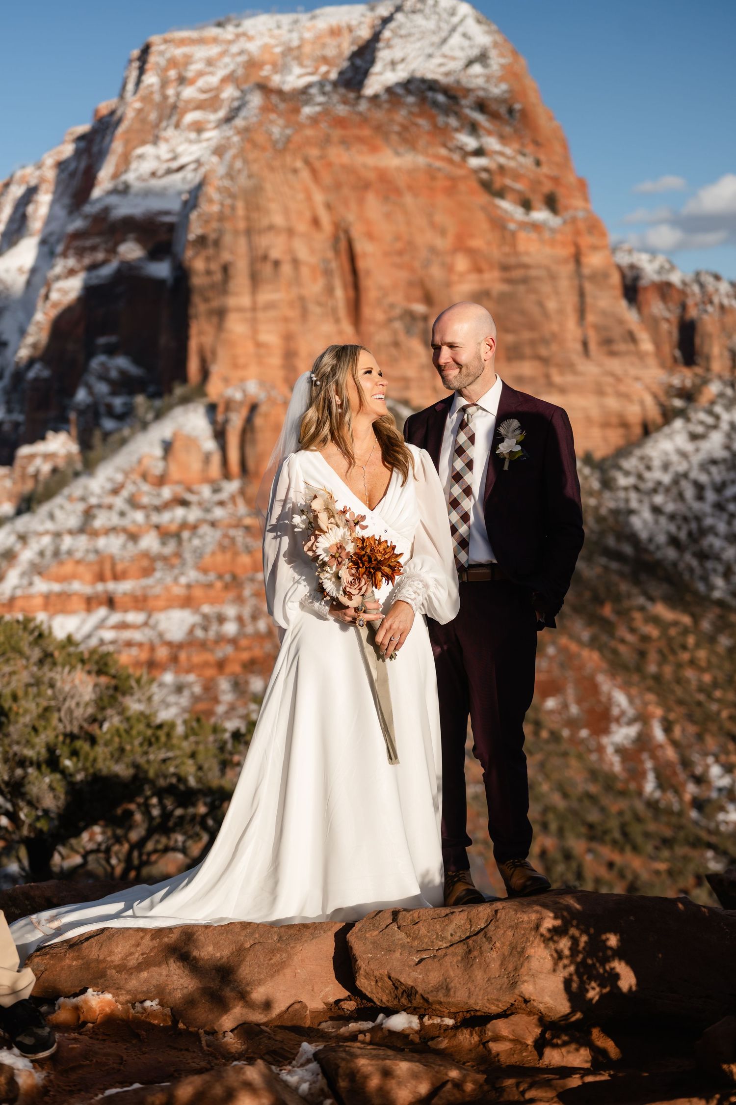 Wedding couple standing on red rock overlook with majestic snow-capped canyon in background.