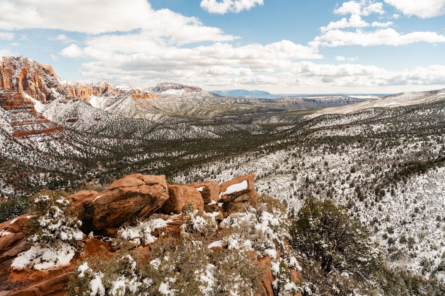 Snow-covered winter landscape with red rock formations and mountains stretching into the distance under blue skies.