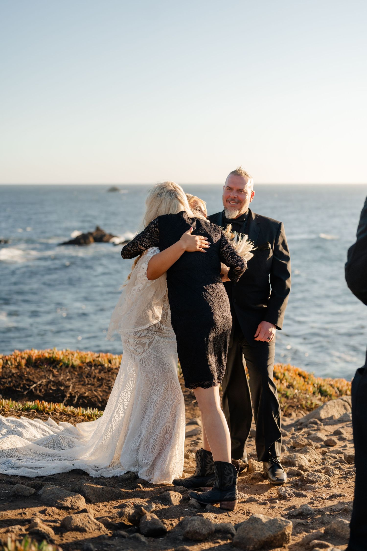 Intimate coastal wedding ceremony taking place on rocky cliffs overlooking the ocean at sunset.