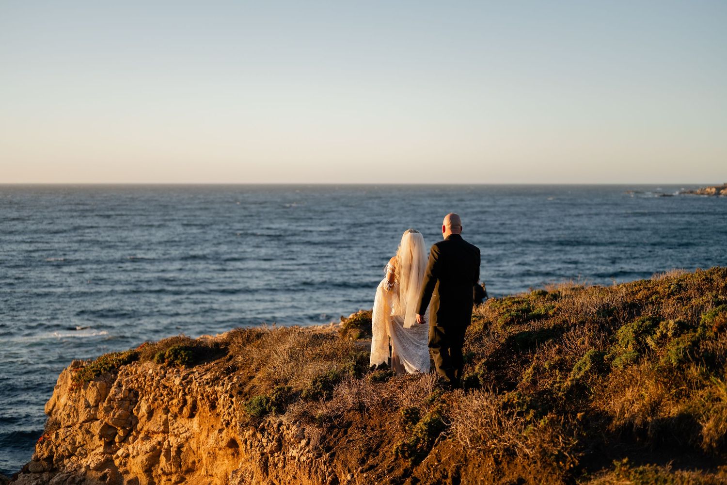 Couple sits on coastal cliffs overlooking the Pacific Ocean during sunset.