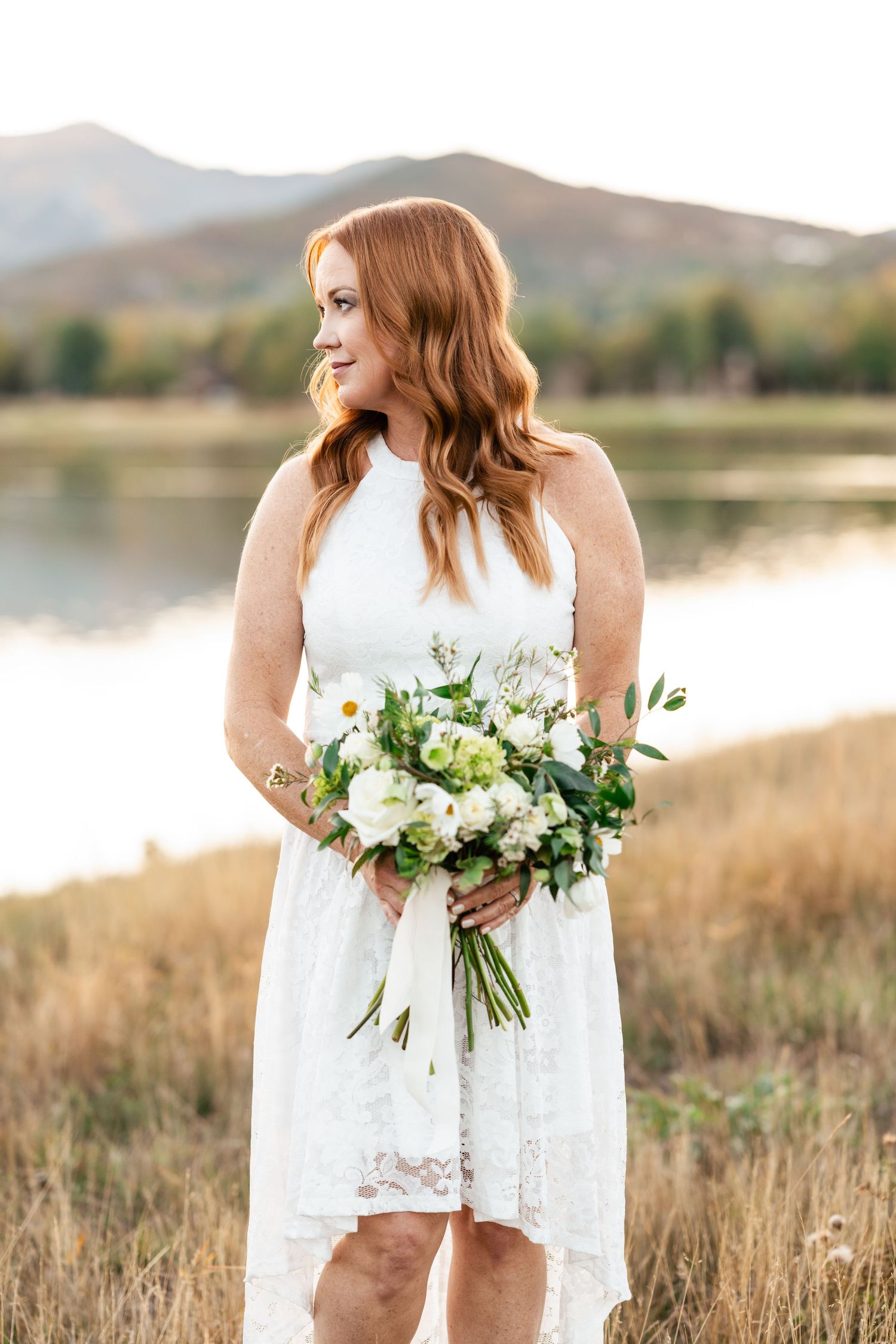 Bride in white lace dress holds elegant white and green bouquet in rustic outdoor setting.