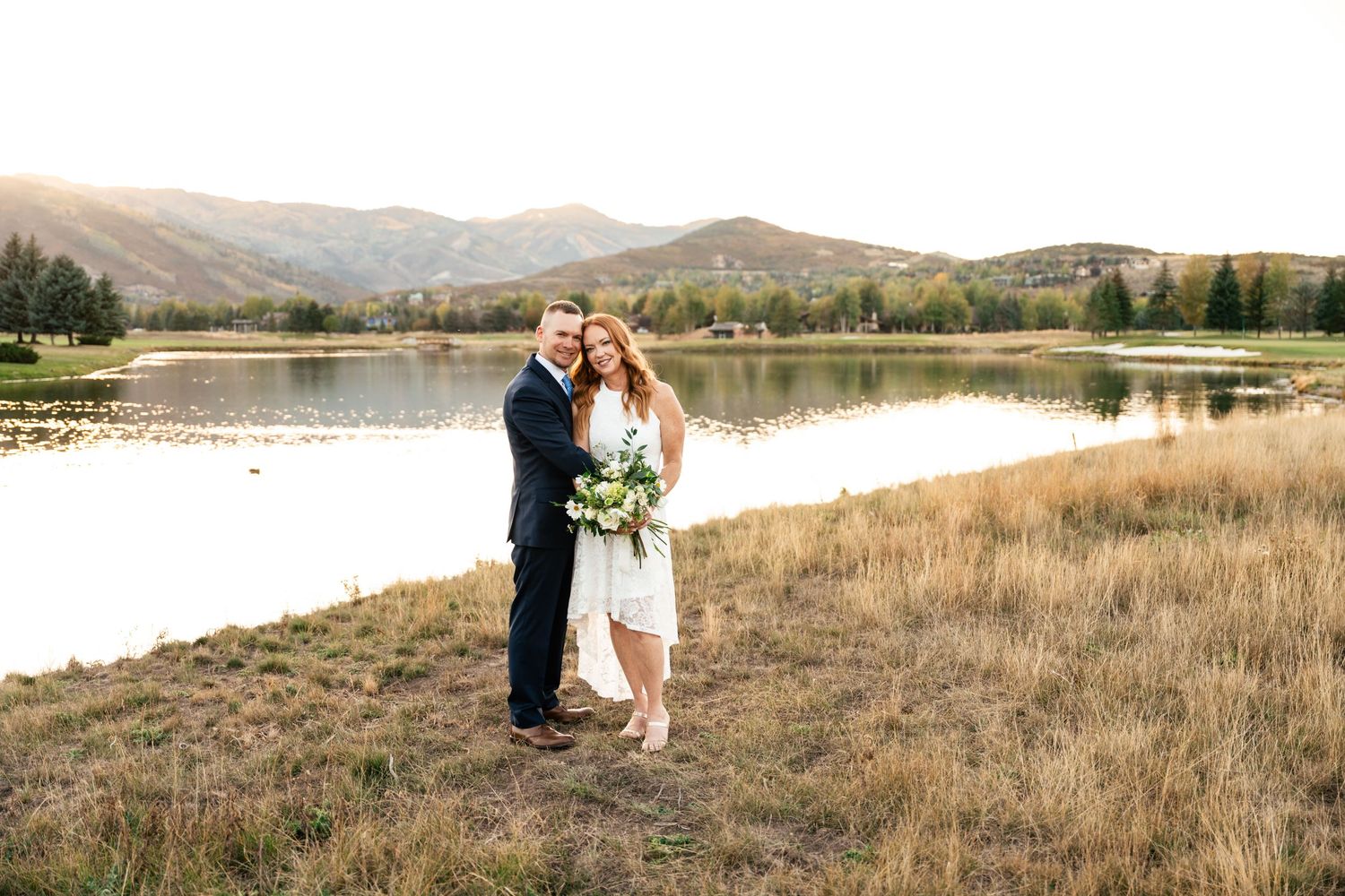 A couple shares a romantic moment by a serene mountain lake surrounded by autumn grass and rolling hills.