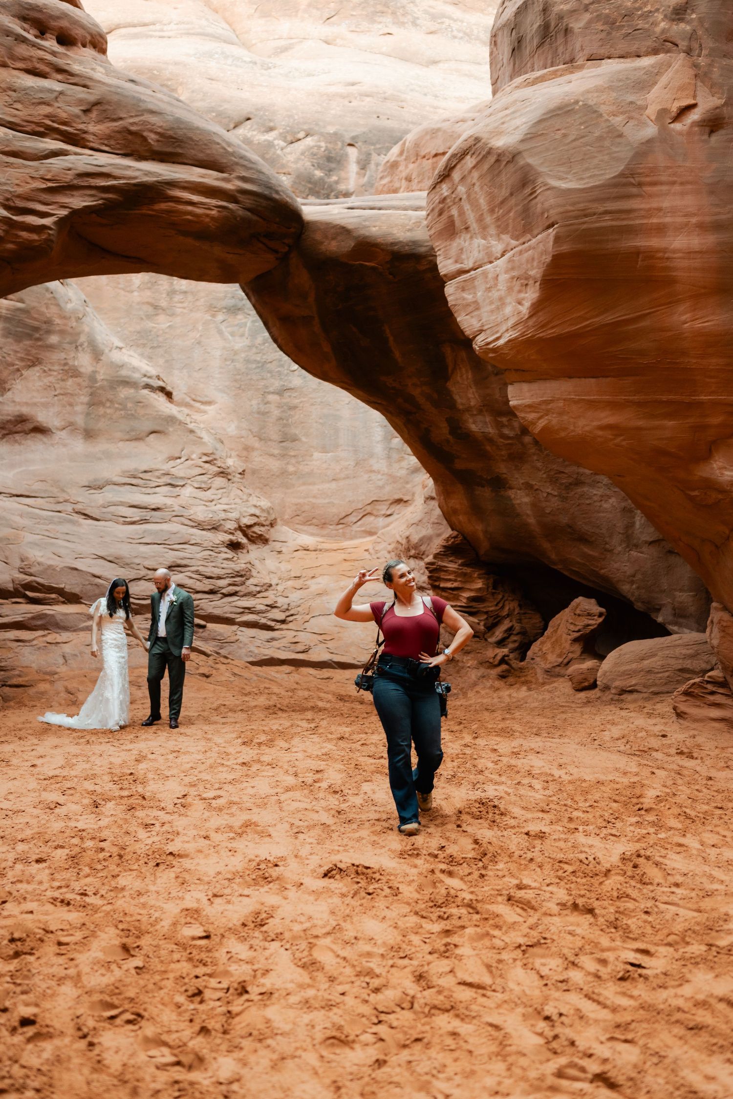 A wedding couple and photographer explore the dramatic red rock arches in a desert landscape.