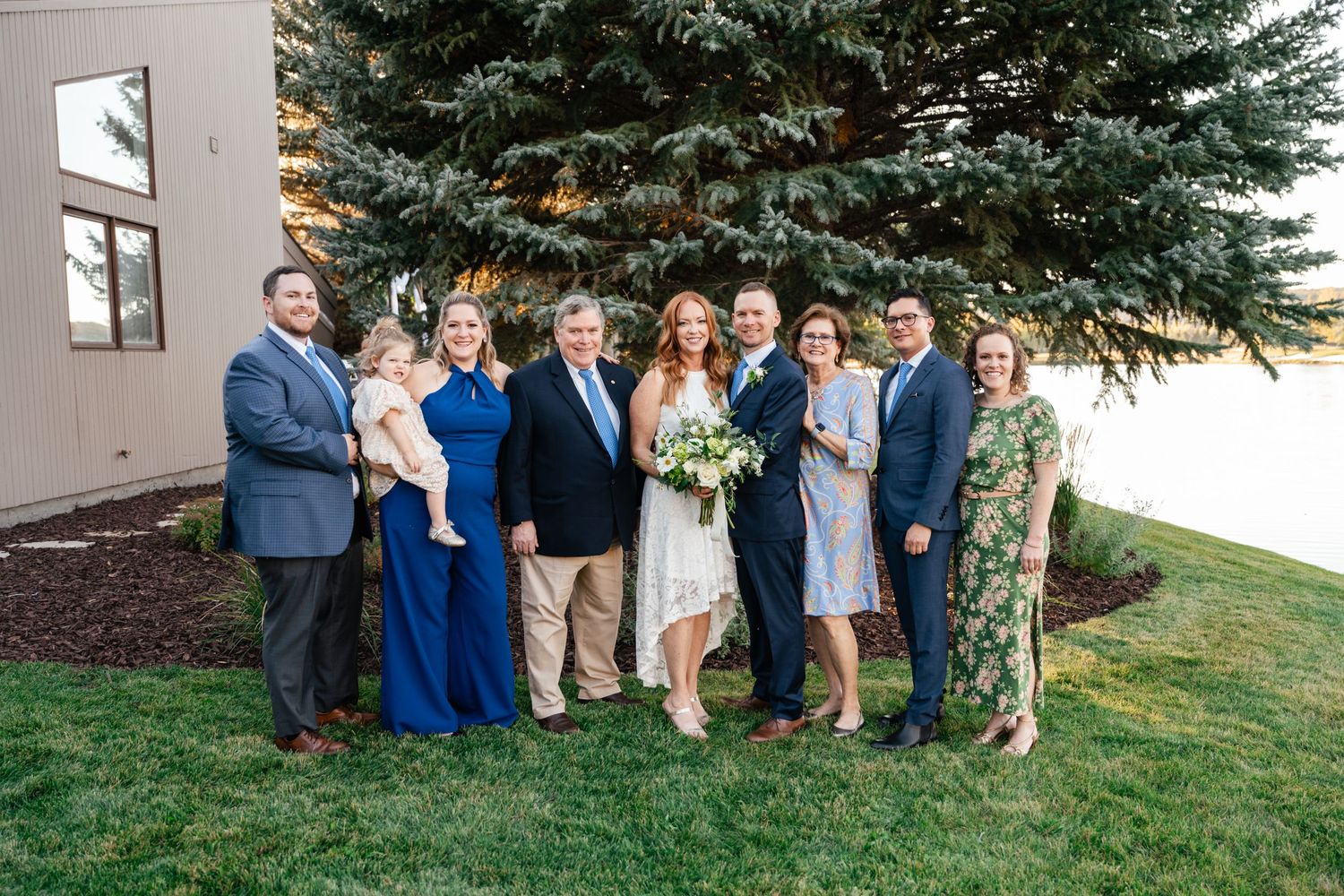 Wedding party gathered outdoors for group photos on a grassy hill near evergreen trees.