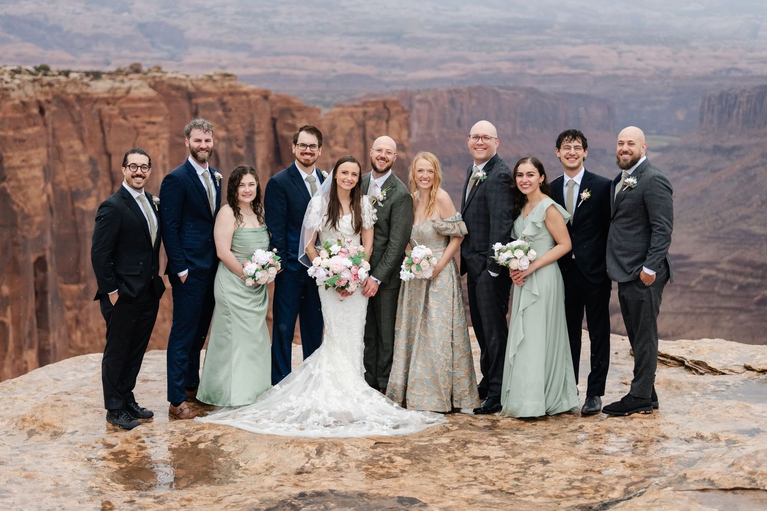 Large wedding group photo with bridal party on scenic desert overlook with canyon views.