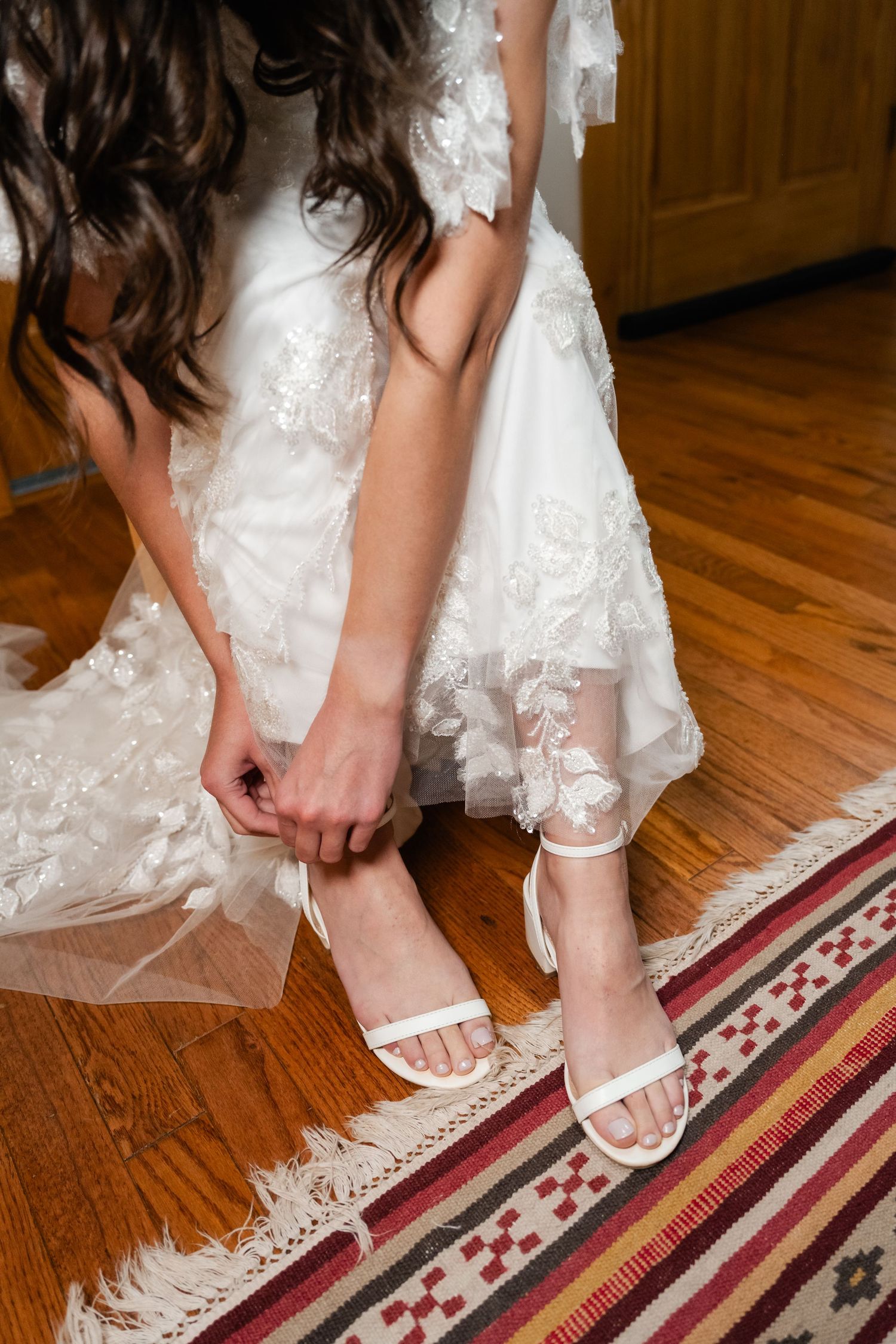 Close-up of bride putting on white sandals while wearing a lace wedding dress on hardwood floor.