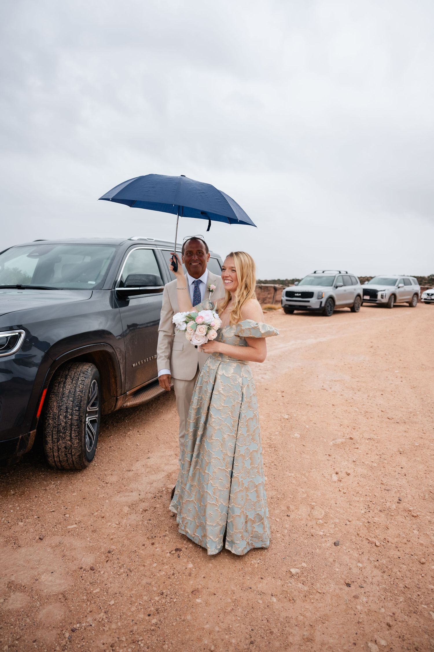 A couple stands by a black car under an umbrella on a desert road during an overcast day.