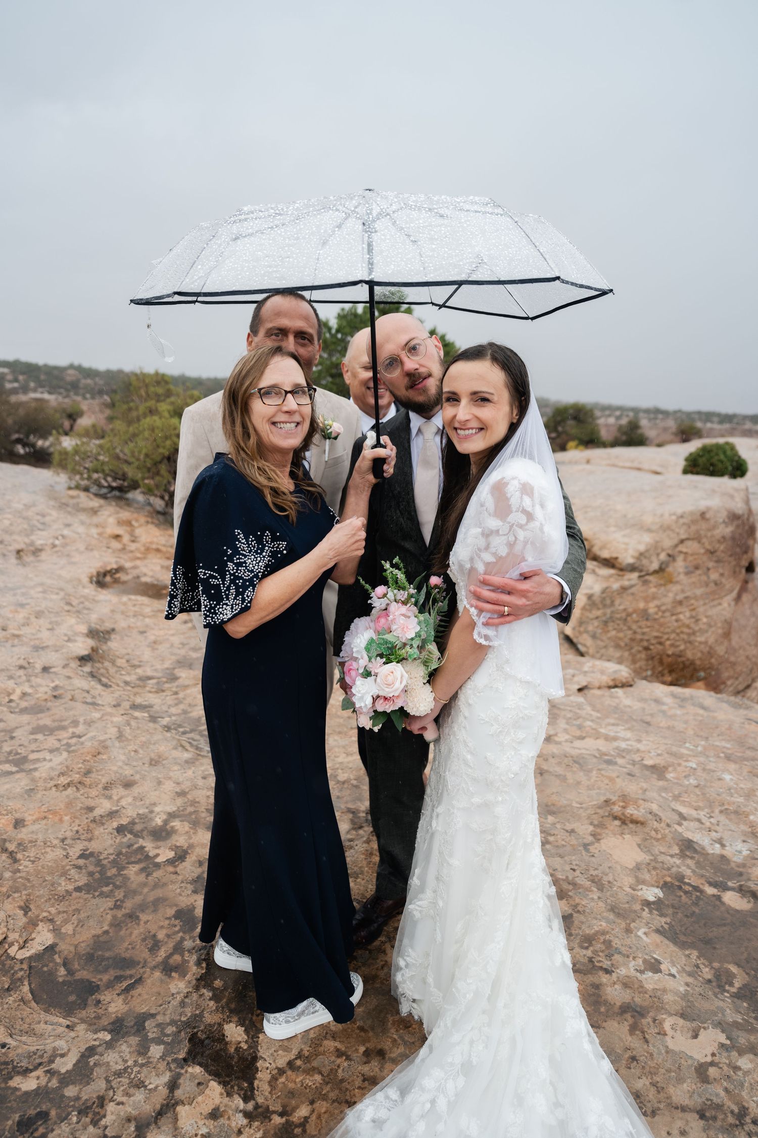 Small wedding group huddled under clear umbrella on rocky outcrop.
