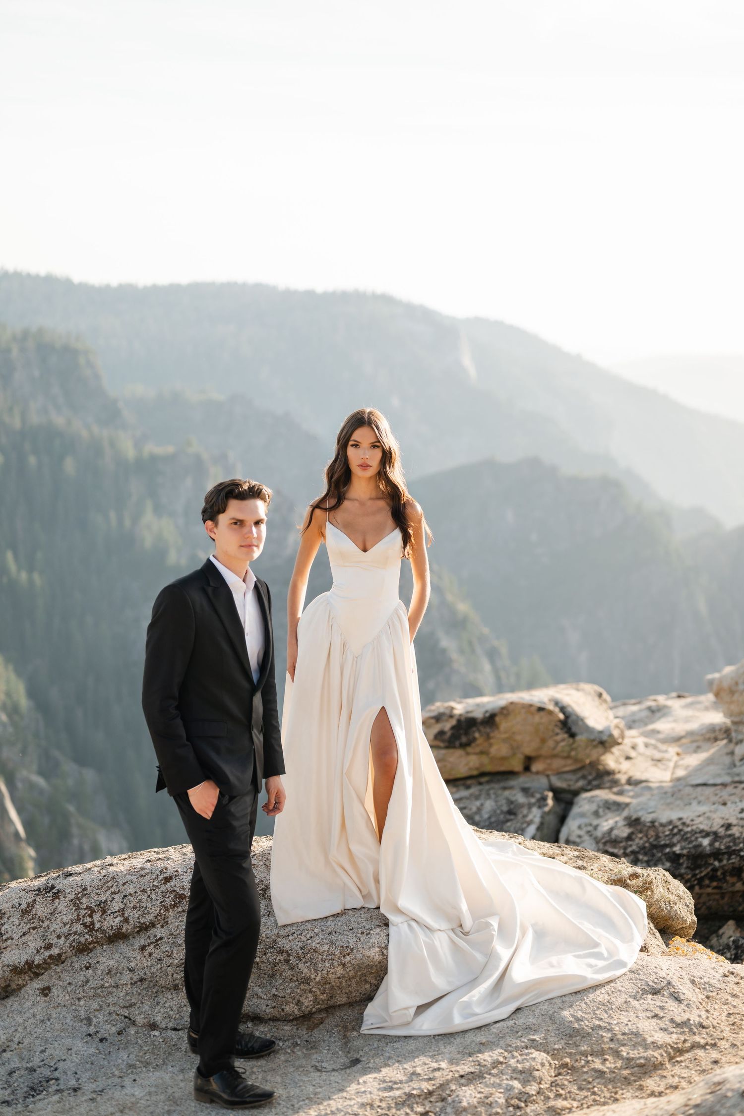 Bride and groom pose on rocky mountain overlook, she in white slit gown, he in black tuxedo.