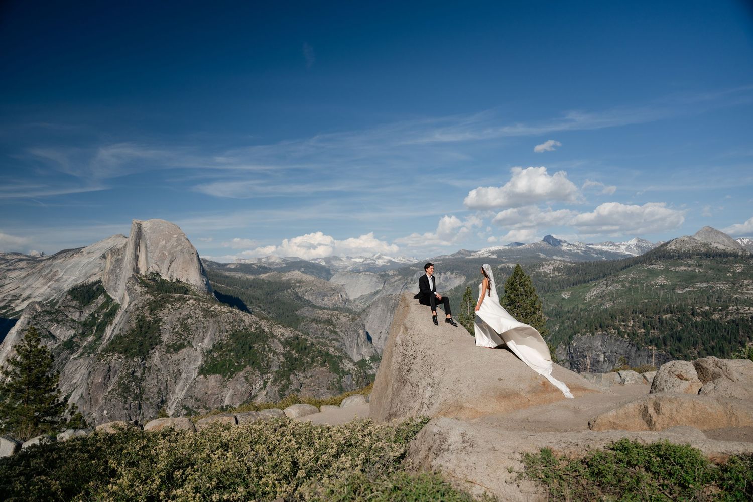 Bride and groom in wedding attire pose on rocky overlook at Yosemite with Half Dome and mountain views.