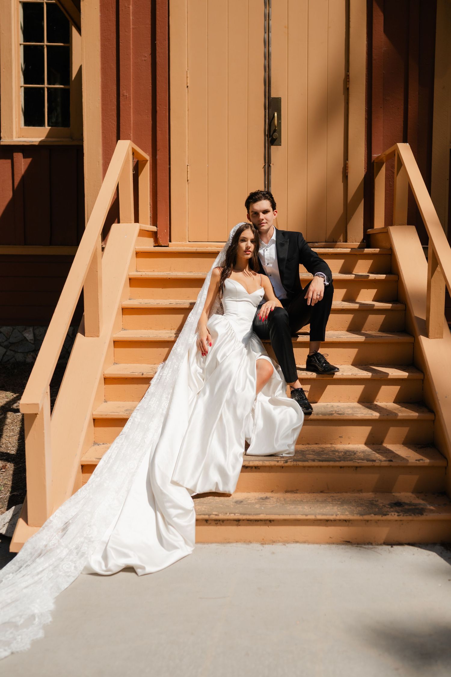 Bride in white gown with veil and groom in black suit sitting on wooden steps outside a rustic building.