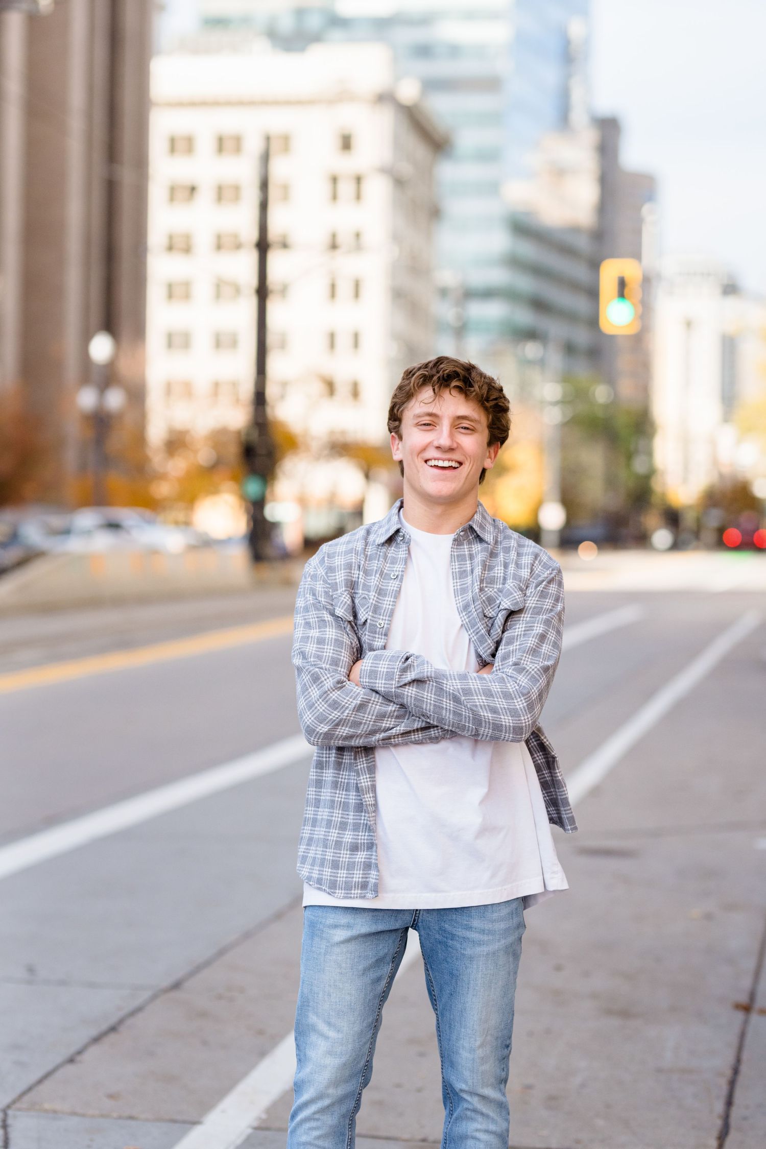 A person wearing a plaid shirt and jeans stands smiling on a city sidewalk with urban buildings in the background.