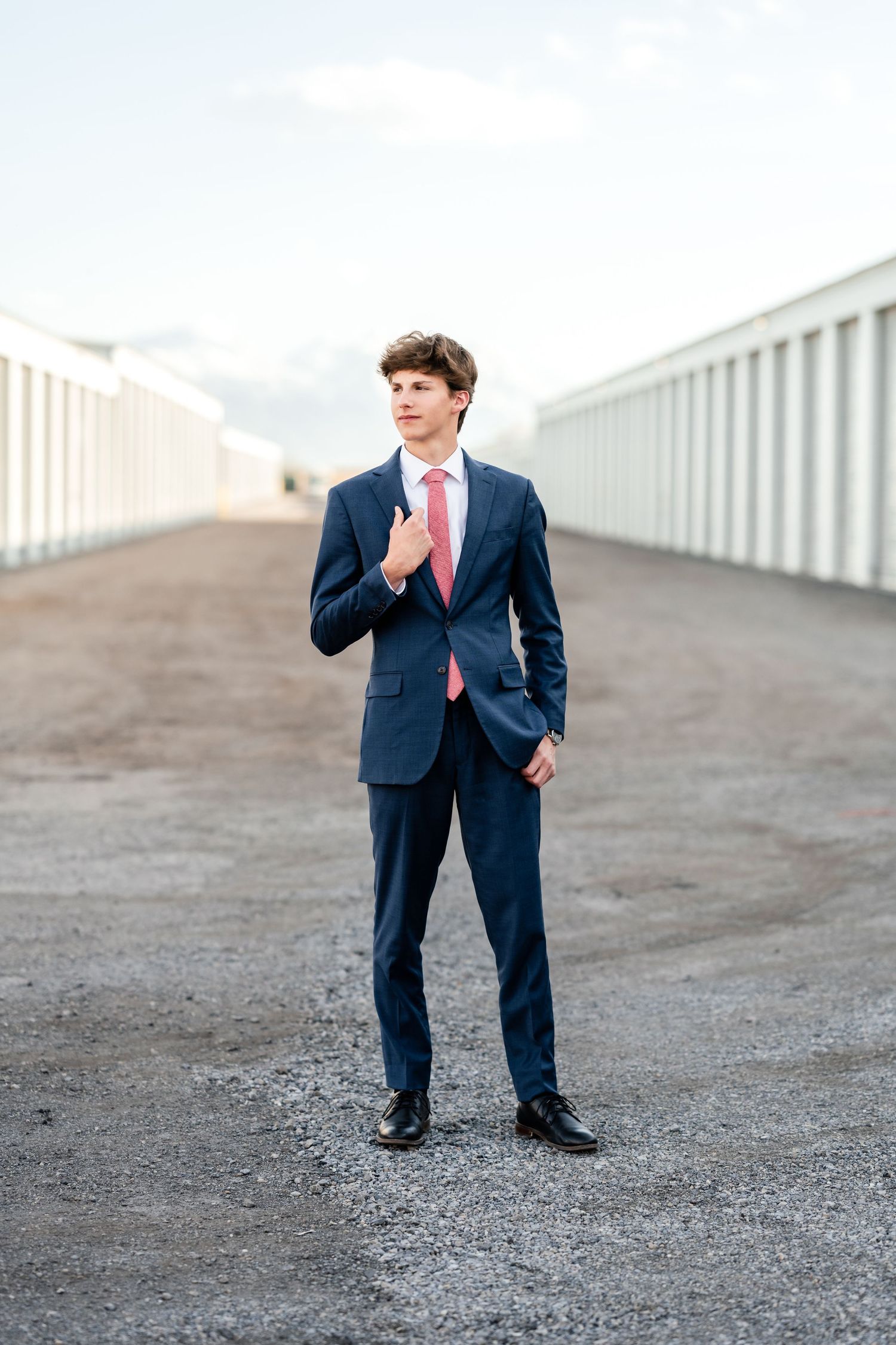 A child in a navy blue suit and pink tie stands on a gravel surface in a professional portrait setting.
