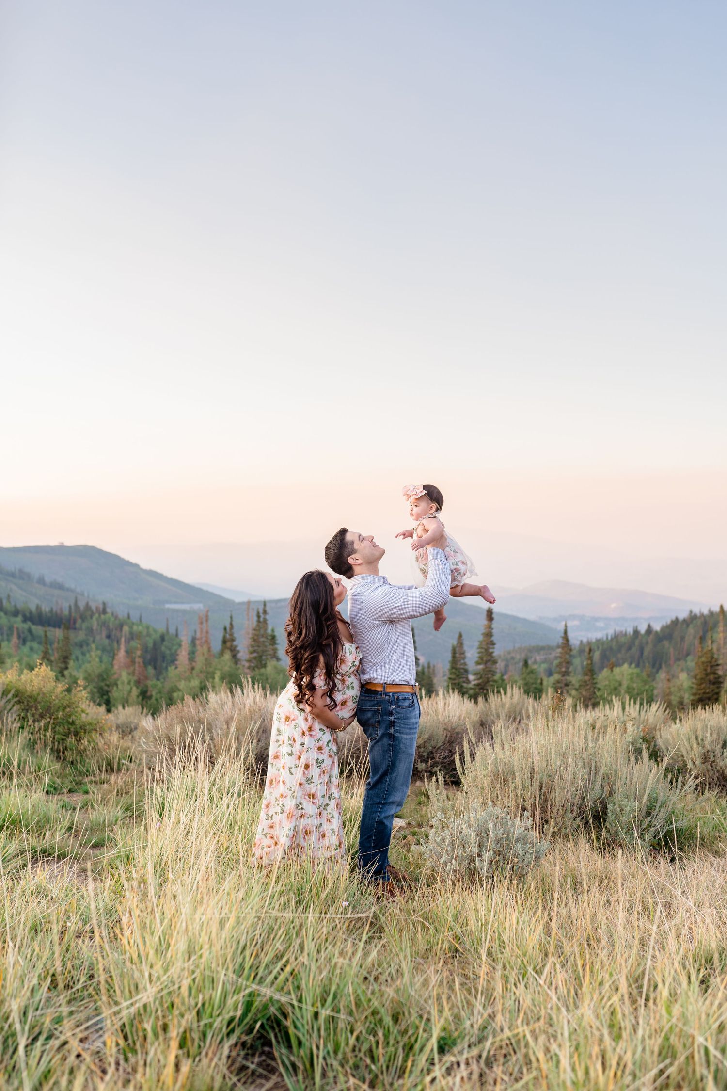 A family stands together in a mountain meadow at sunset with rolling hills and forest in the background.