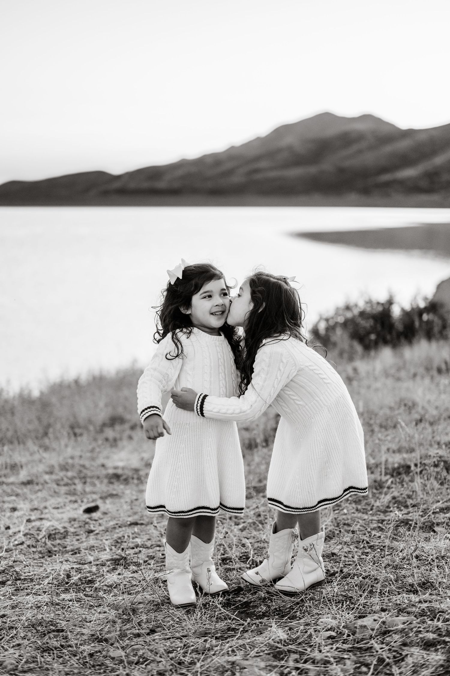 Two young children in white dresses share a tender moment by a scenic lake with mountains in the background.