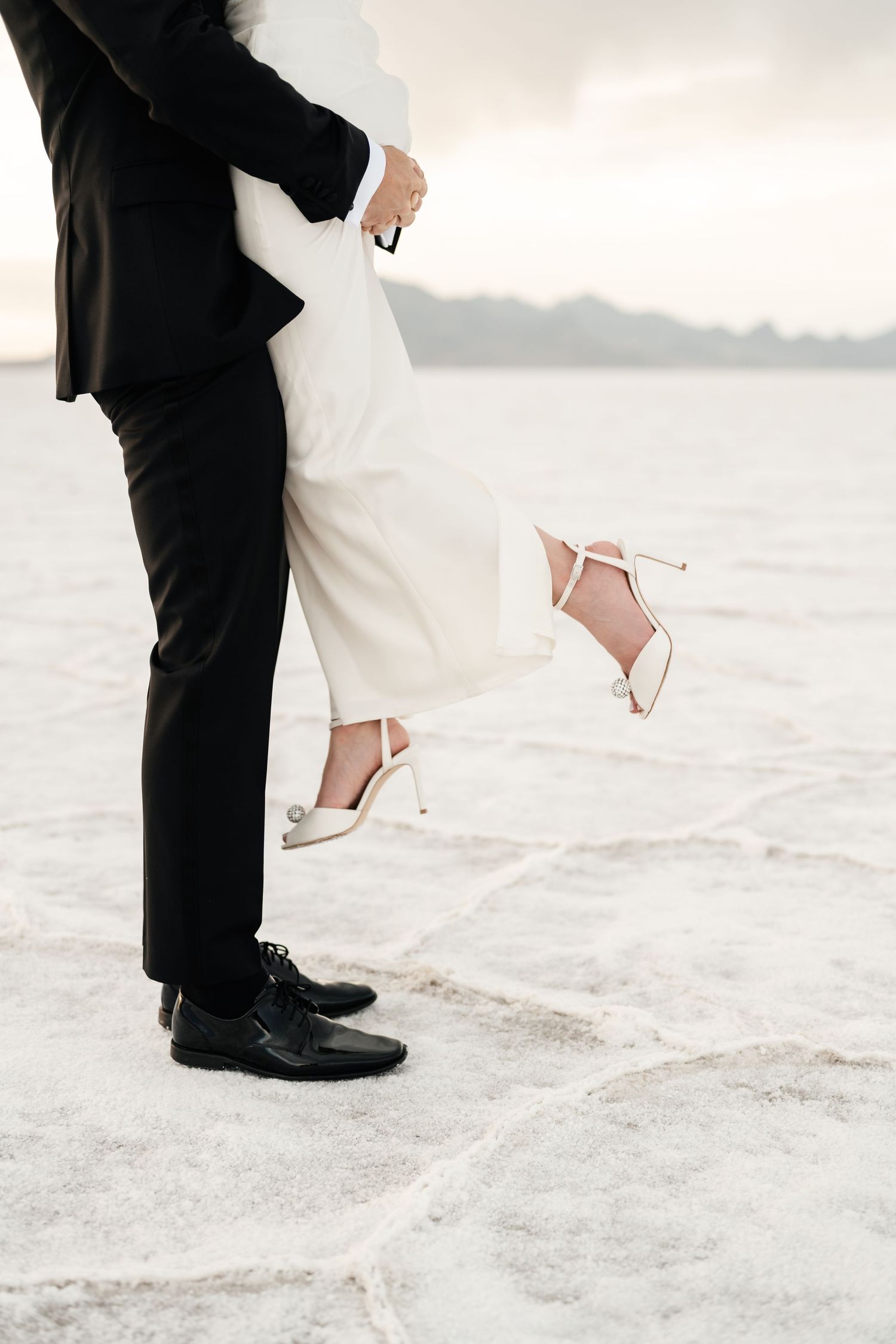 Salt Flats Elopement Photographer. Close-up view of formal shoes and wedding dress hem on white salt flats during sunset.