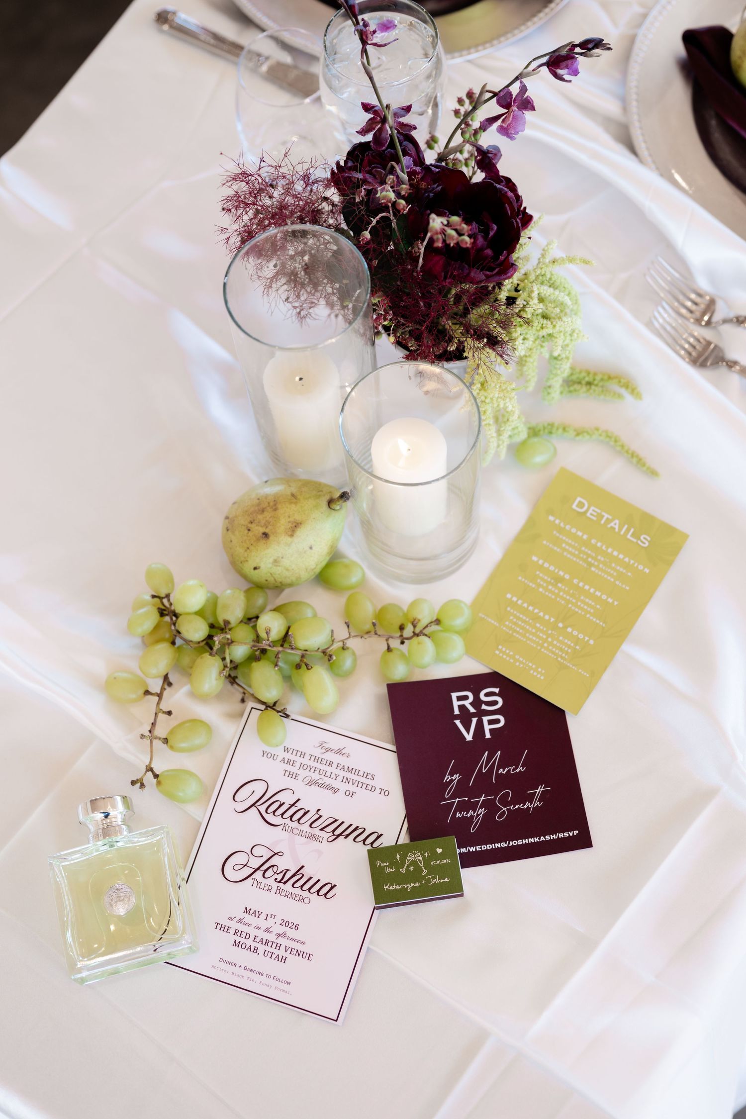 Elegant wedding table with invitation suite, RSVP card, candles, green grapes, and burgundy floral centerpiece on white linen.