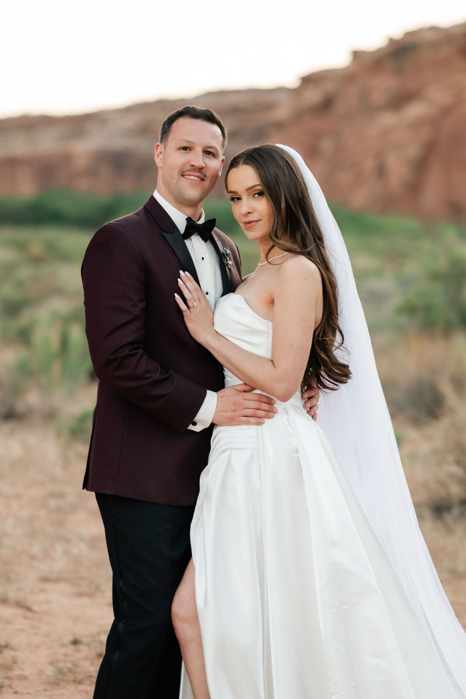 Bride and groom embracing in desert landscape, groom in burgundy tuxedo, bride in white strapless gown with veil.