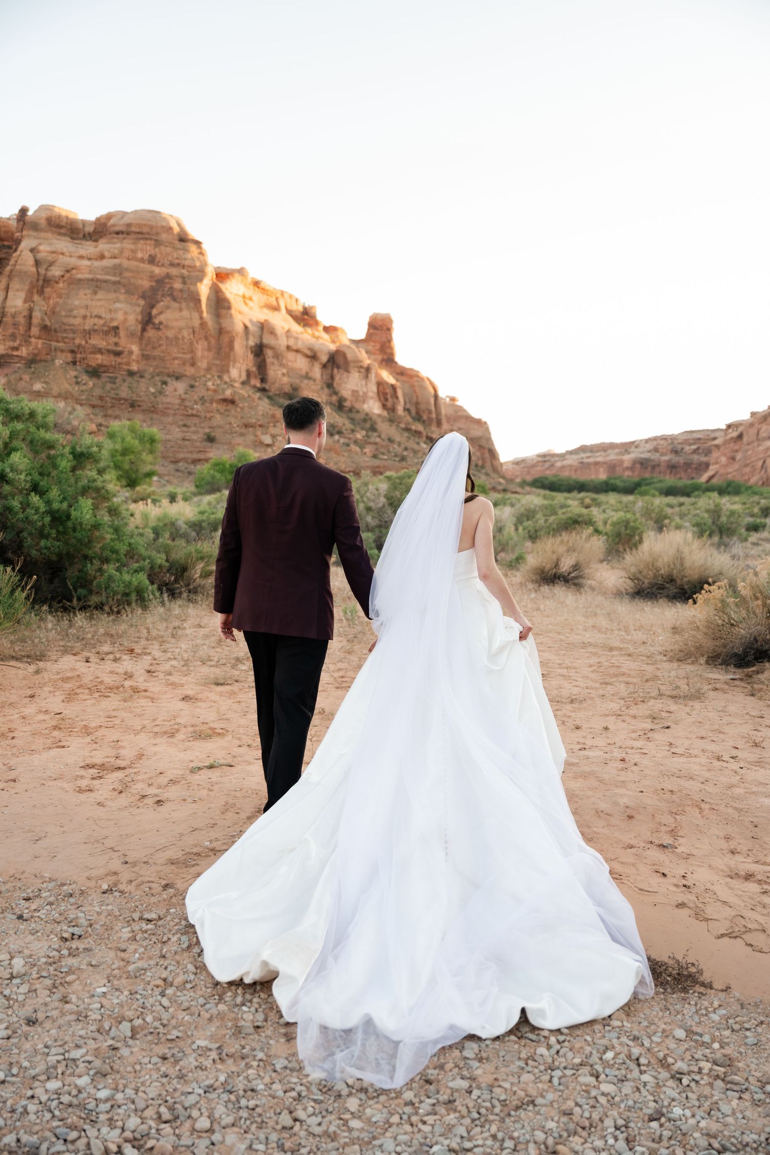 Bride and groom walk hand in hand away from camera toward towering red rock formations in desert landscape.