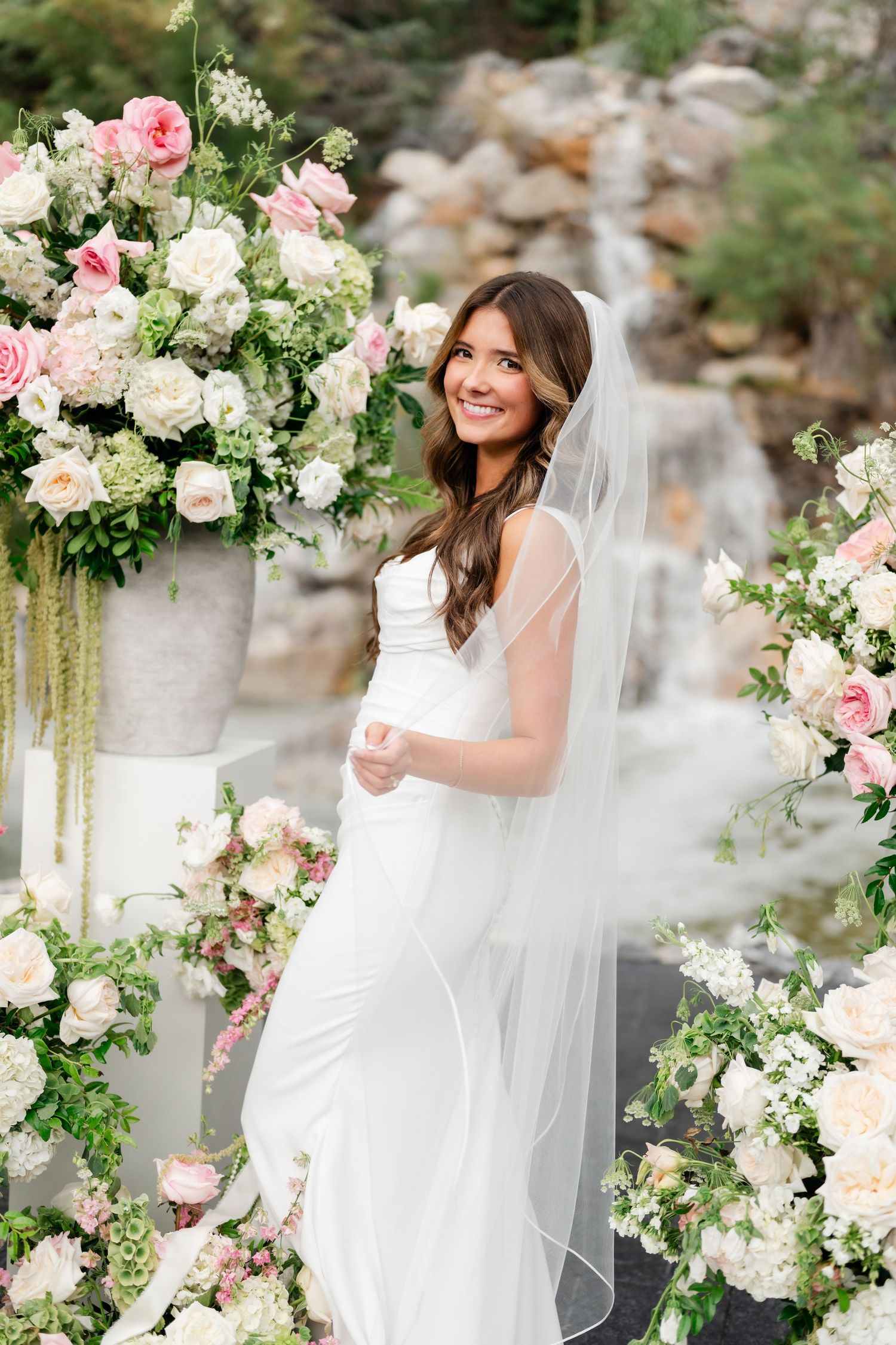 Smiling bride in white gown and veil surrounded by lush pink and white floral arrangements near a waterfall backdrop.