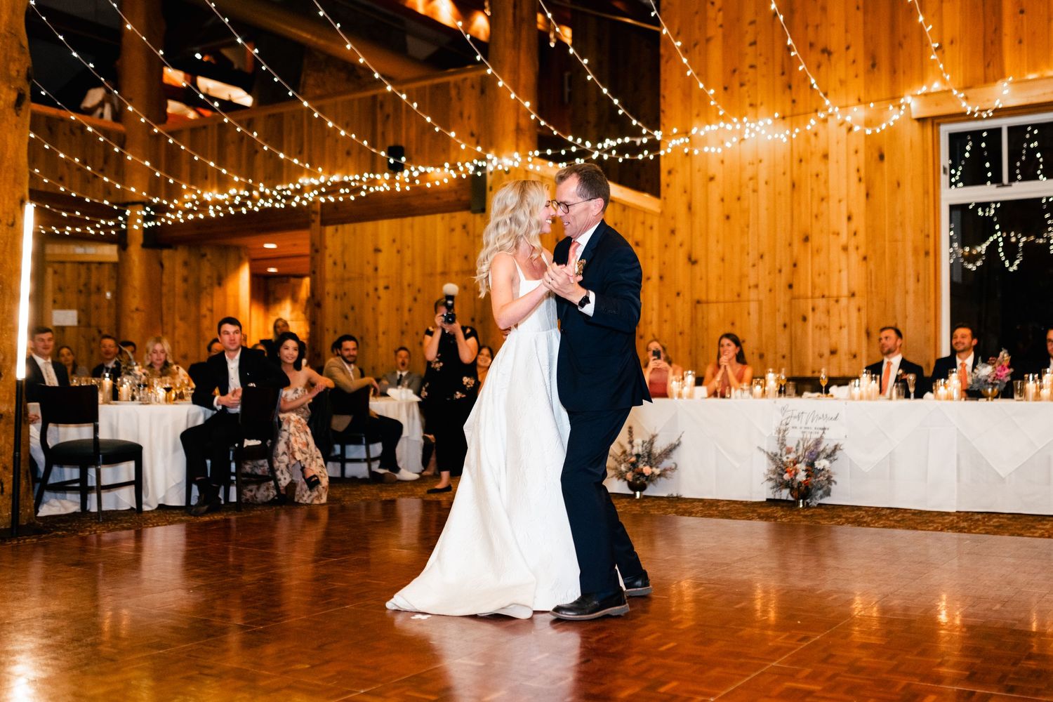 A couple shares their first dance in a rustic barn venue decorated with twinkling string lights and white reception tables.