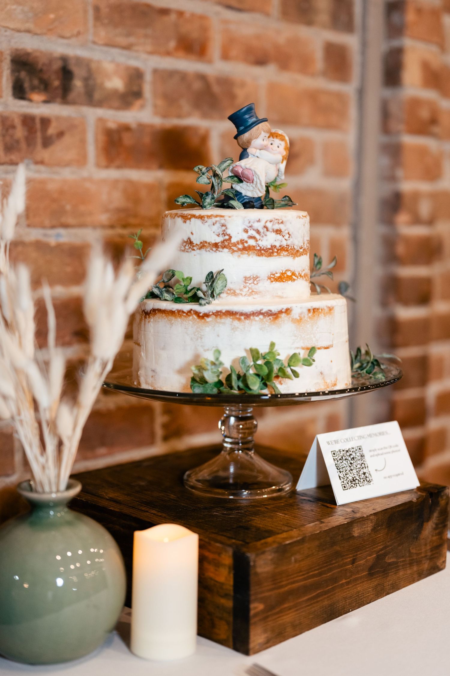 A semi-naked wedding cake decorated with eucalyptus greenery and white buttercream sits on a glass pedestal near a brick wall.