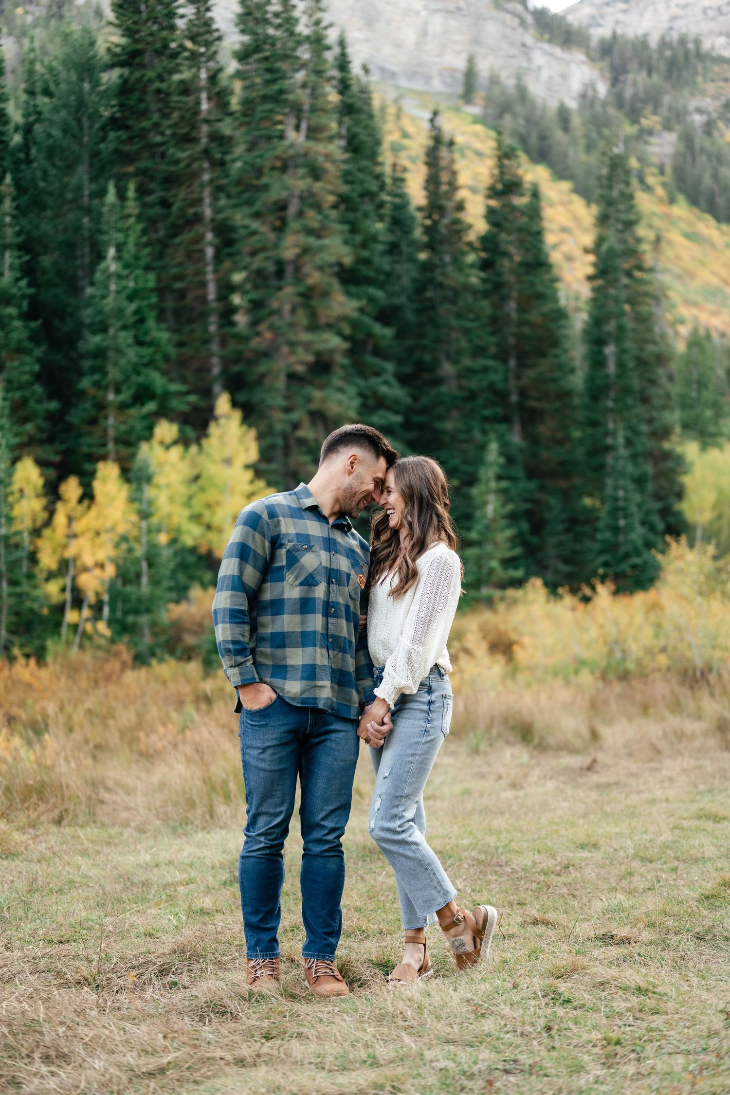 A couple shares an intimate moment in a mountain meadow with evergreen trees and peaks behind them.