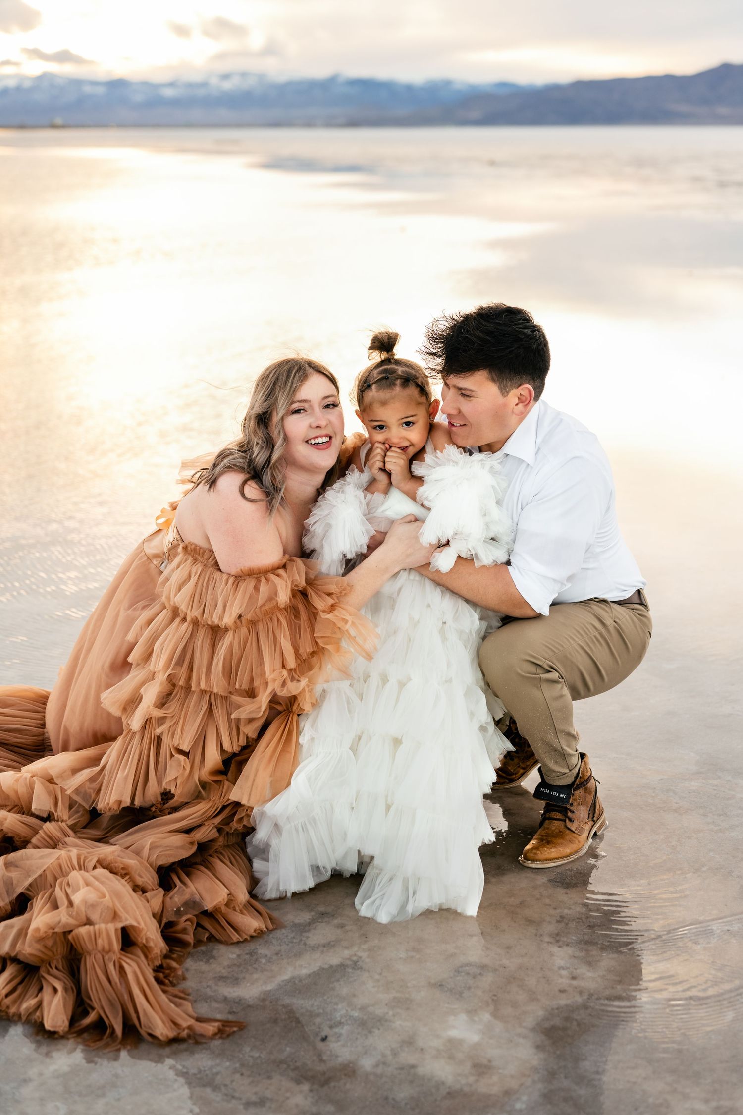 Parents embrace their child affectionately amidst the vast expanse of the flooded salt flats during golden hour.