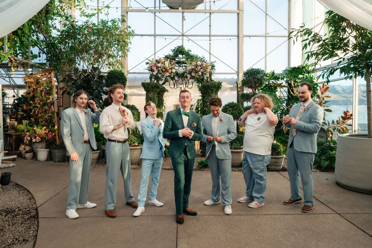 Group of groomsmen in casual suits and sweaters posing together in a bright greenhouse setting with florals.
