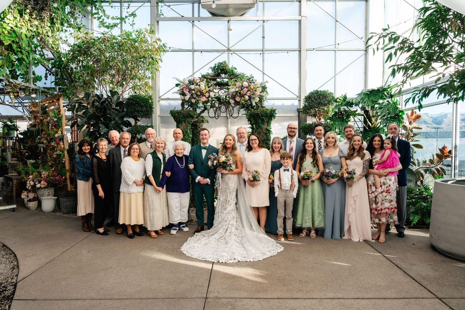 Wedding party group photo in a bright greenhouse with floral decor and natural lighting.
