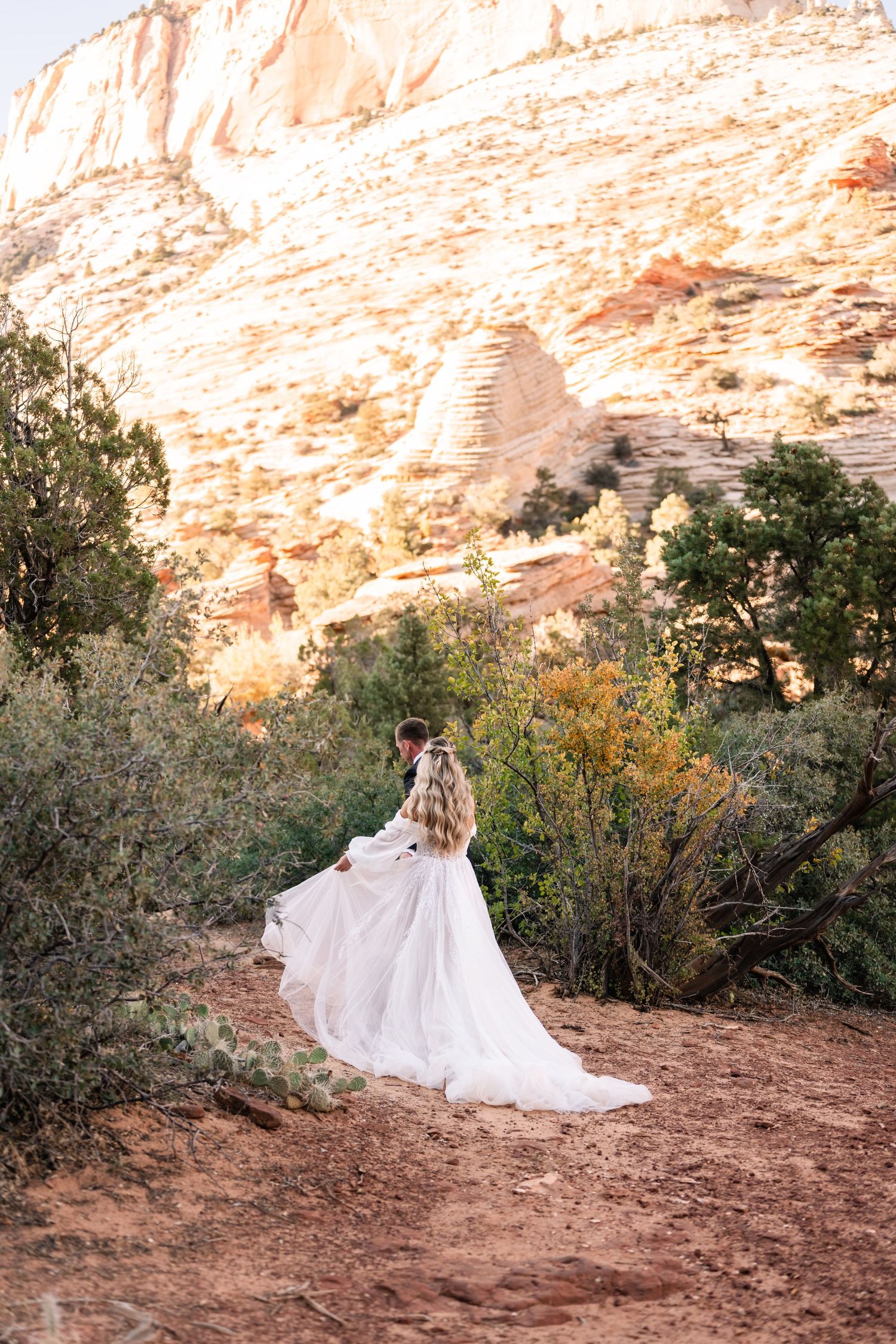 Bride in flowing white gown stands alone against majestic desert canyon backdrop.