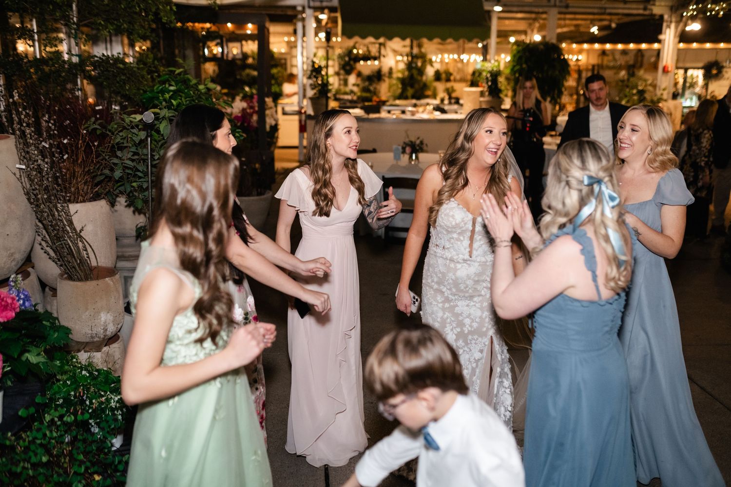 Wedding reception guests dancing and laughing together at an evening celebration with decorative lights and greenery.